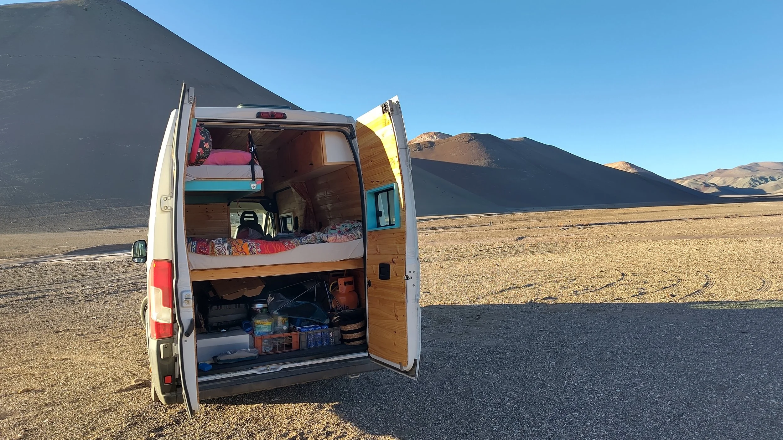 Ein weißer Campingbus mit geöffnetem Seitenteil, das eine Inneneinrichtung mit Bett, Stauraum und Küchenbereich zeigt, steht in einer Trockensalzwüste mit Bergen im Hintergrund, bei klarem blauen Himmel.