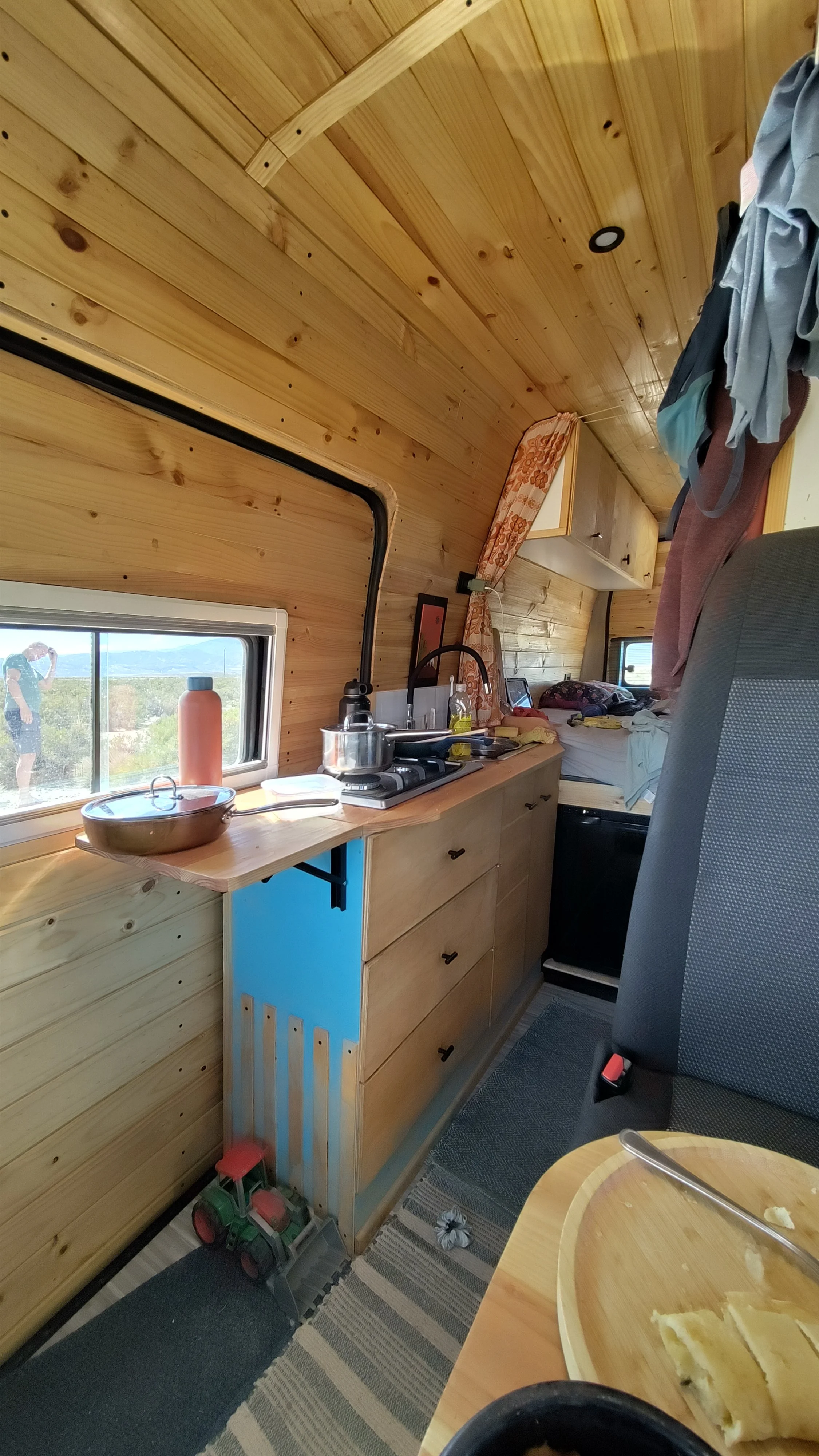 Innenansicht eines kleinen Holzwohnmobils mit Mini-Küche, einem Bett im Hintergrund, einer Person im Spiegel, und einigen Spielsachen auf dem Boden.