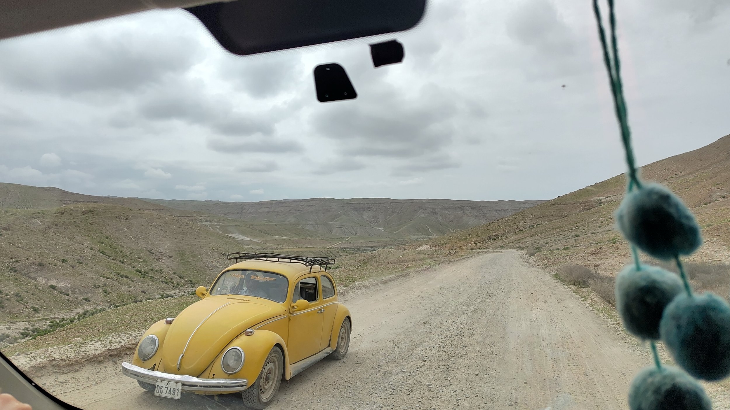 Ein gelber, vintage Volkswagen Käfer fährt auf einer unbefestigten, staubigen Landstraße durch eine karge, bergige Landschaft. Der Blick ist aus einem Fahrzeug, das wahrscheinlich im Inneren oder am Armaturenbrett aufgenommen wurde, inklusive einer b