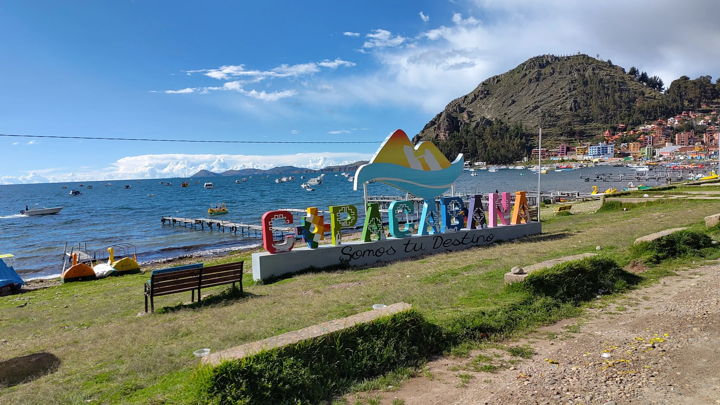 Blick auf den See mit zahlreichen Booten, im Vordergrund ein bunter Willkommensschild mit dem Text "COPACANA" und der Aufschrift "Somos tu Destino". Im Hintergrund sind Berge und eine bunte Stadt. Der Himmel ist teilweise bewölkt.