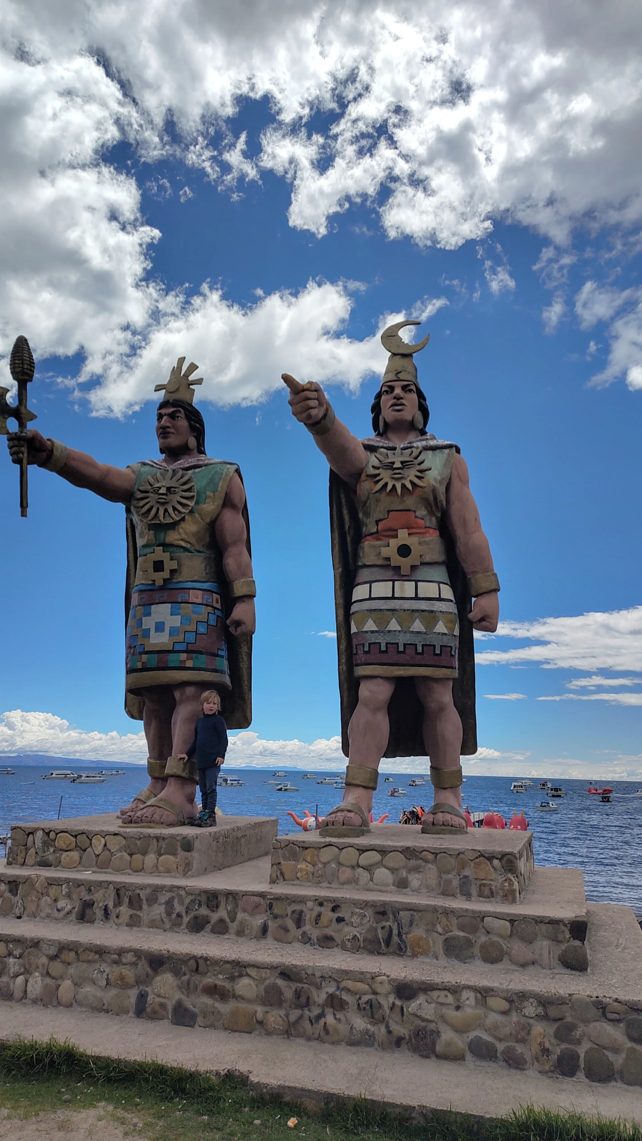 Zwei große Statuen von ehemaligen Kriegern vor einer Wasserfront mit Booten, eine kleine Person steht zwischen ihnen auf einer Steinplattform, der Himmel ist mit Wolken bedeckt.