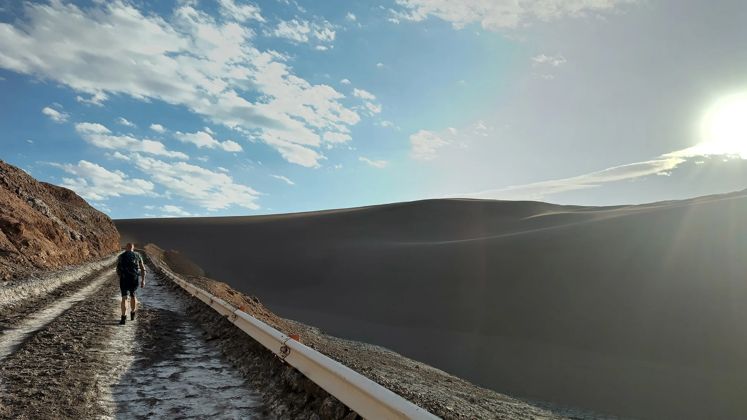 Ein Wanderer läuft auf einem matschigen Pfad neben einer langen, weißen Schutzbarriere durch eine Wüstenlandschaft mit großen Sanddünen, bei strahlendem Himmel und Sonne.