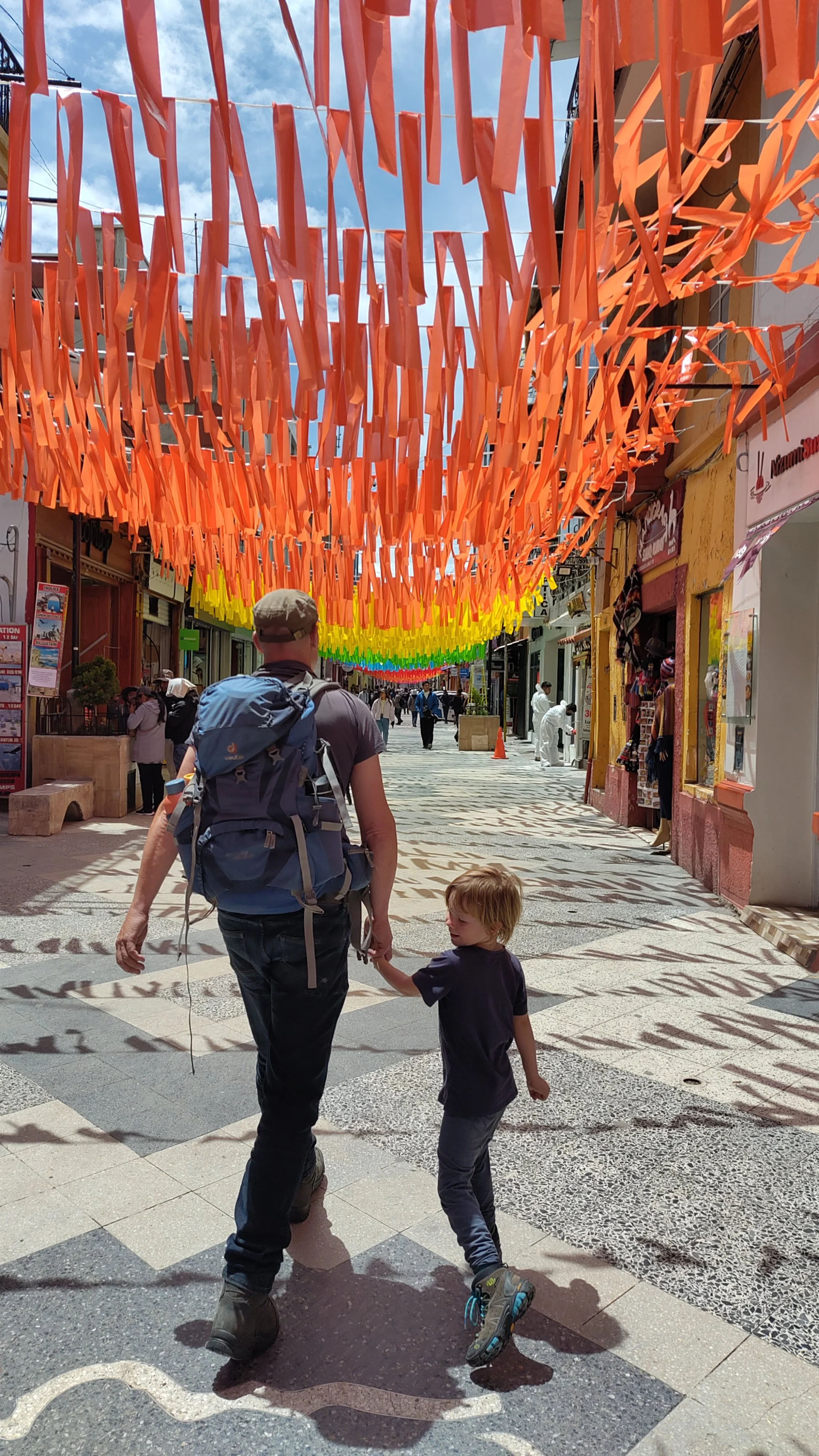 Straße mit bunten Papierstreifen in Orange, Gelb, Grün und Blau, Menschen, darunter ein Mann mit einem Rucksack und ein Kind, im Hintergrund Geschäfte und Passanten.