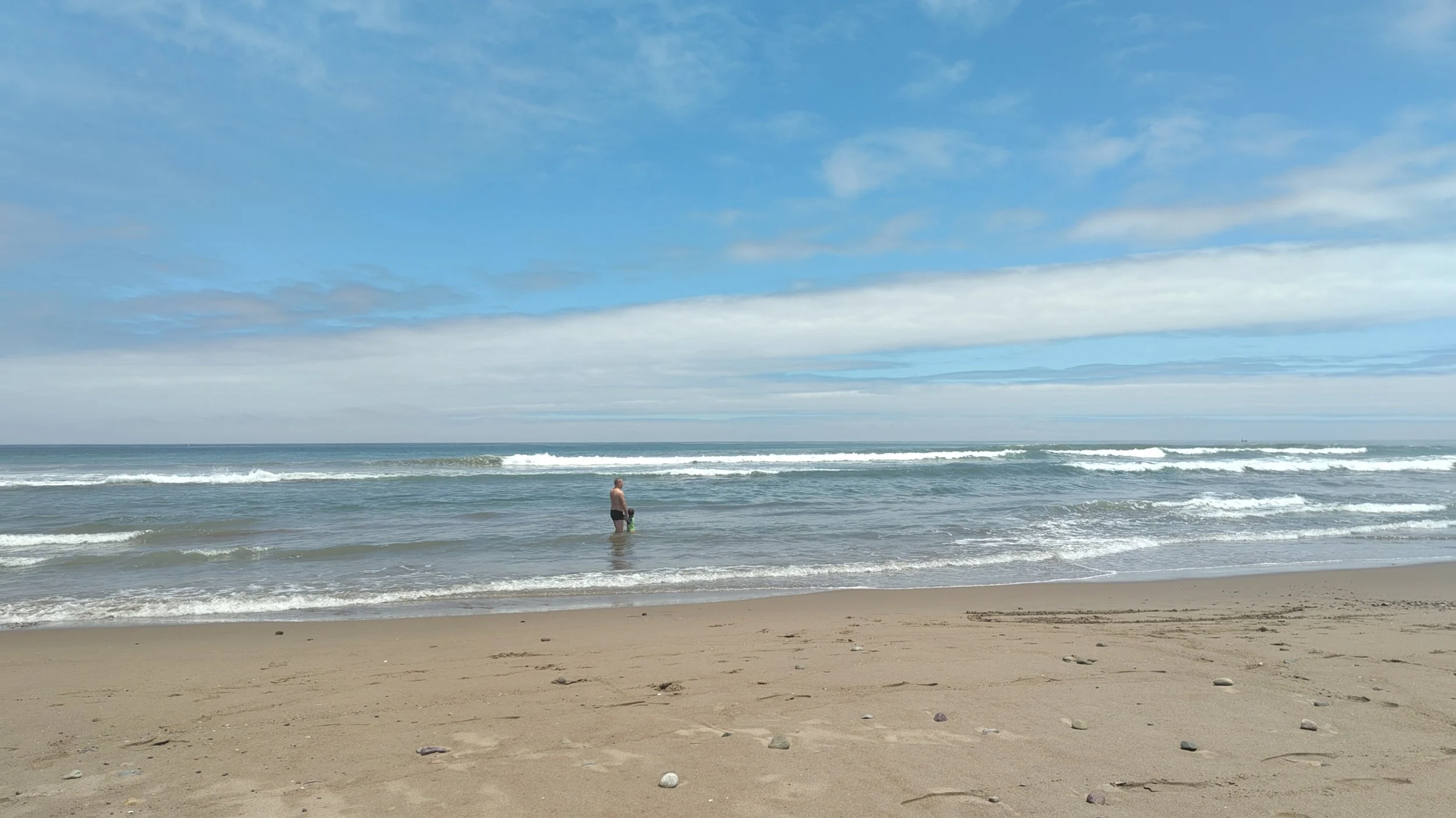 Ein einzelner Mann steht im Wasser am Strand, blickt aufs Meer, bei bewölktem Himmel.