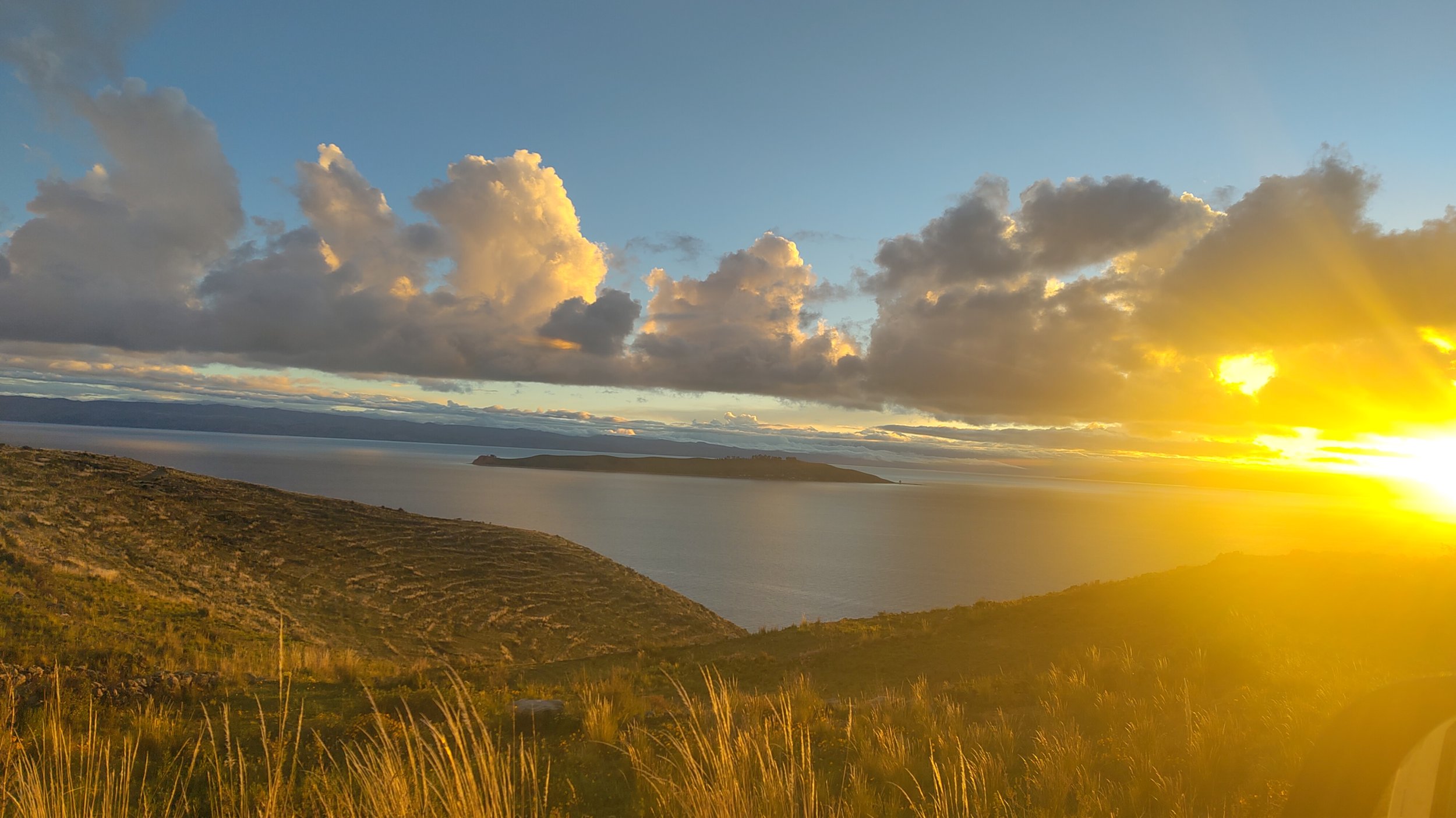 Blick auf eine Bucht bei Sonnenuntergang mit bewölktem Himmel und hügeliger Landschaft im Vordergrund.