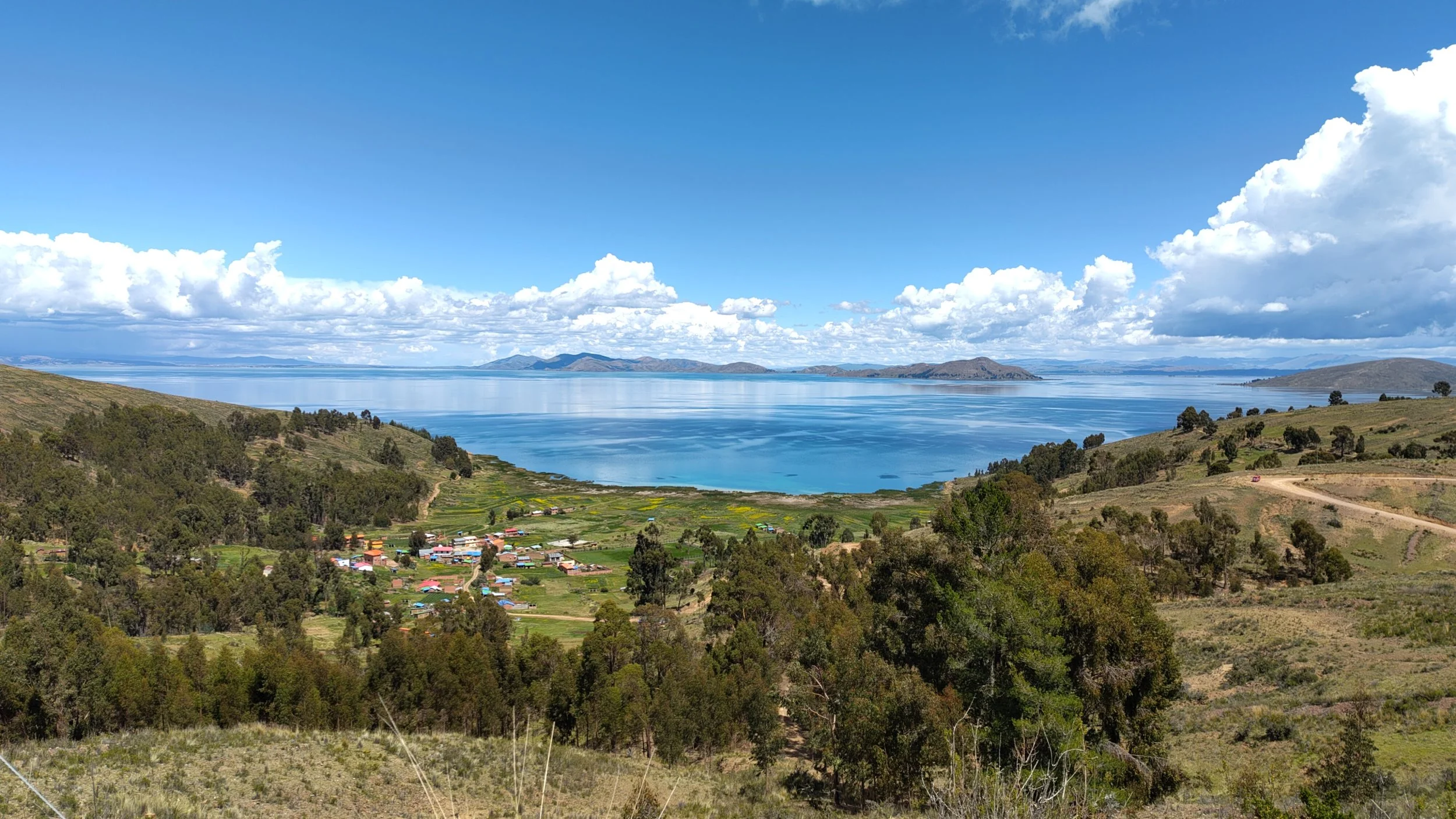 Großer See mit Hügeln im Hintergrund, bewölkter Himmel, kleine Dorfsiedlung im Vordergrund, umgeben von grünen Wäldern und Feldern.
