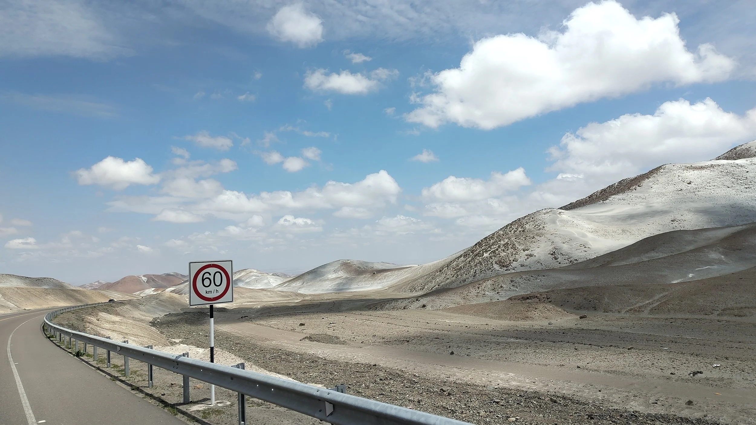 Straße mit Geschwindigkeitsbegrenzung auf 60 km/h, umgeben von kargen, gewölbten Hügeln unter einem Himmel mit einigen Wolken.
