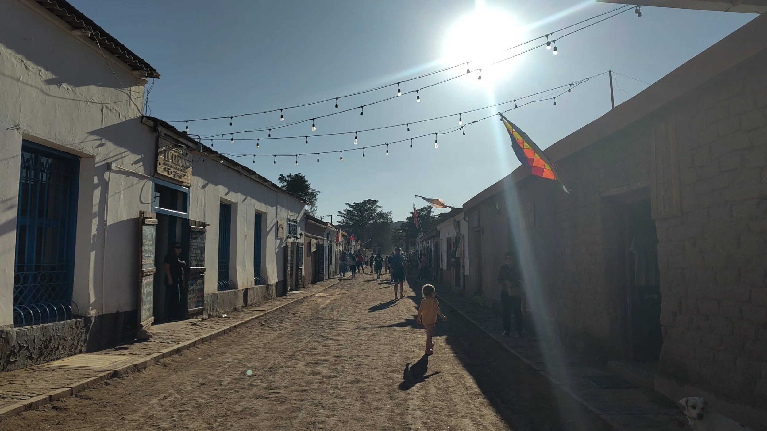 Straße in einem kleinen Ort mit weißen Gebäuden, Menschen laufen, Sonnenschein und bunte Flaggen. Stromleitungen sind sichtbar.