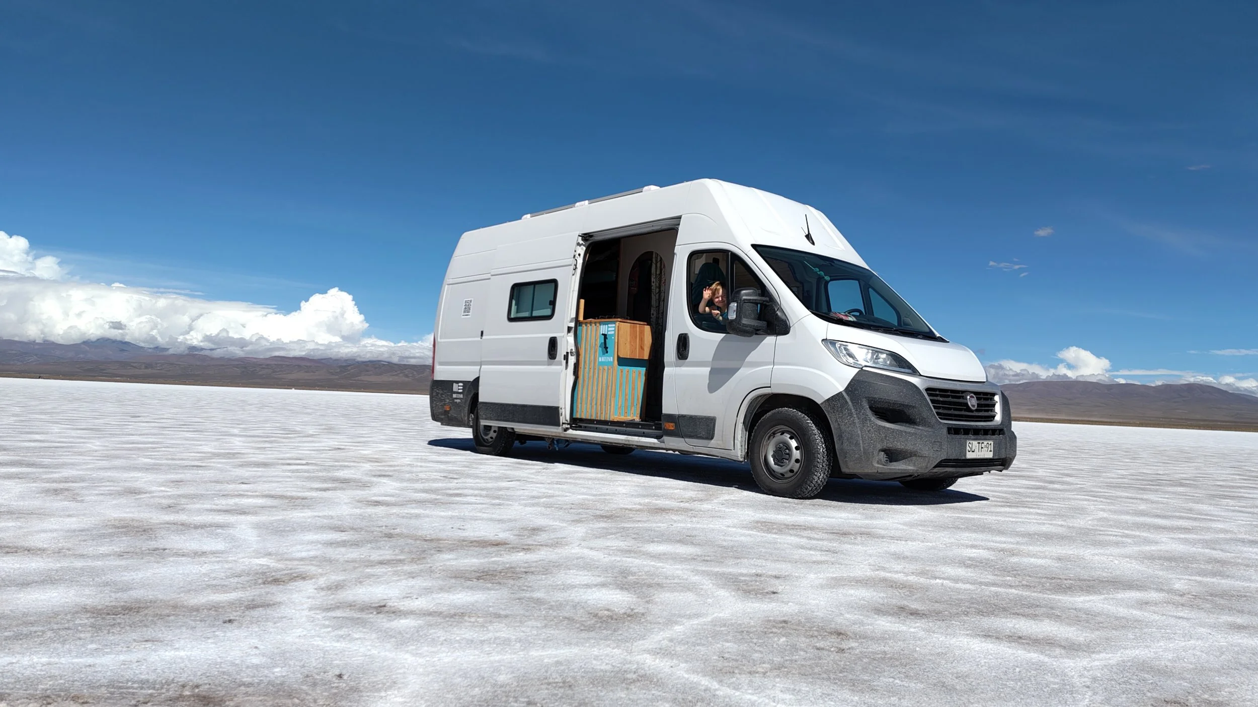 Ein weißer Camper auf einer weiten, trockenen, flachen Landschaft mit Bergketten im Hintergrund und einer blauen Himmel mit wenigen Wolken