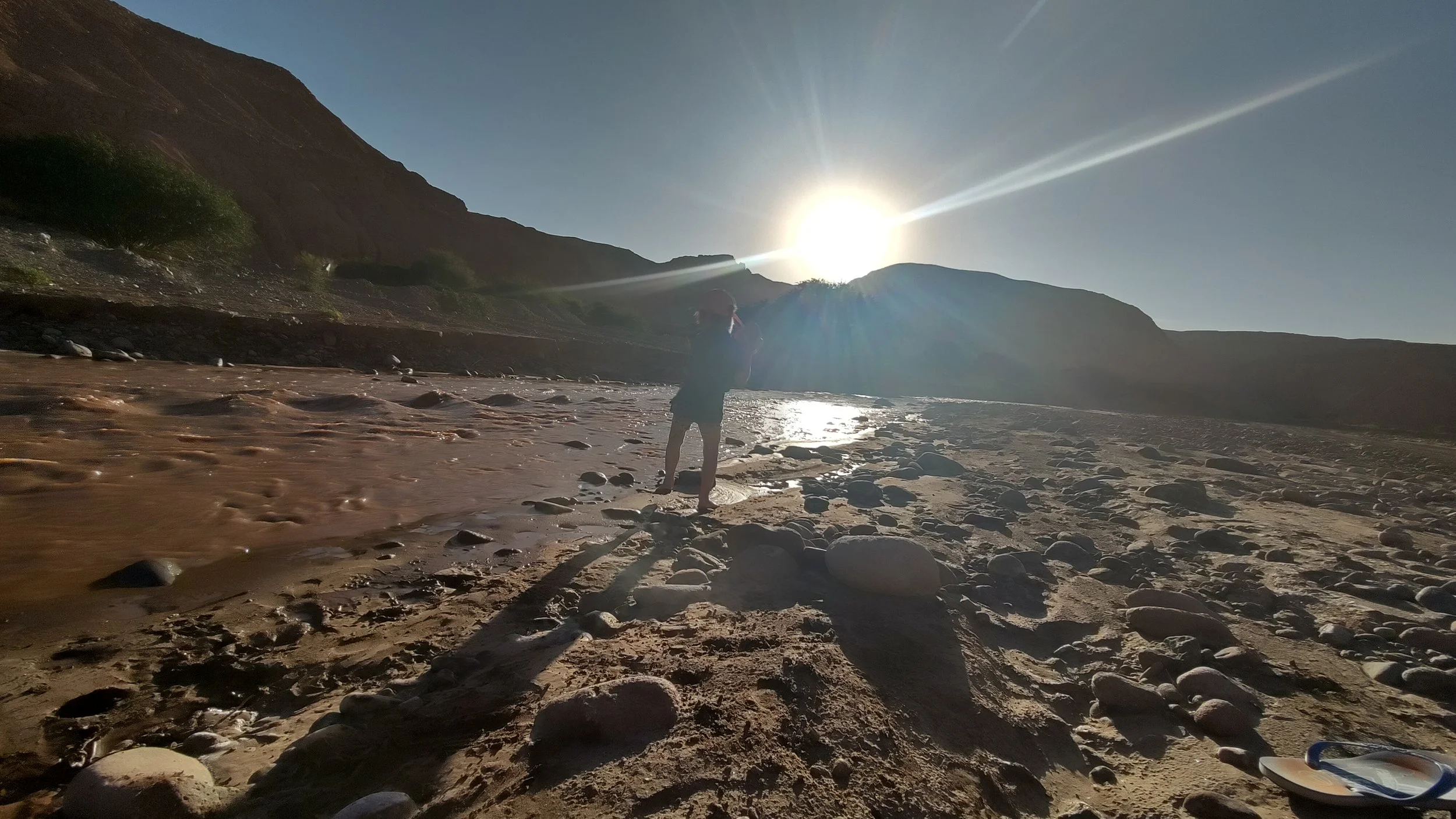 Person steht am Strand bei Sonnenuntergang, umgeben von Bergen, mit Sonnenstrahlen am Himmel.