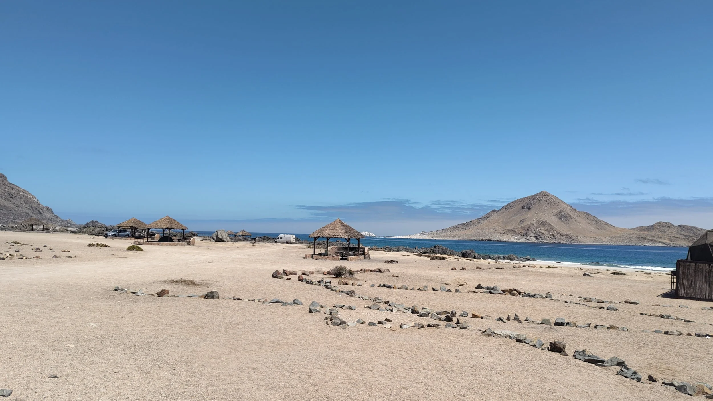 Strand mit Sonnenschirmen, Fahrzeugen und Bergen im Hintergrund bei blauem Himmel.