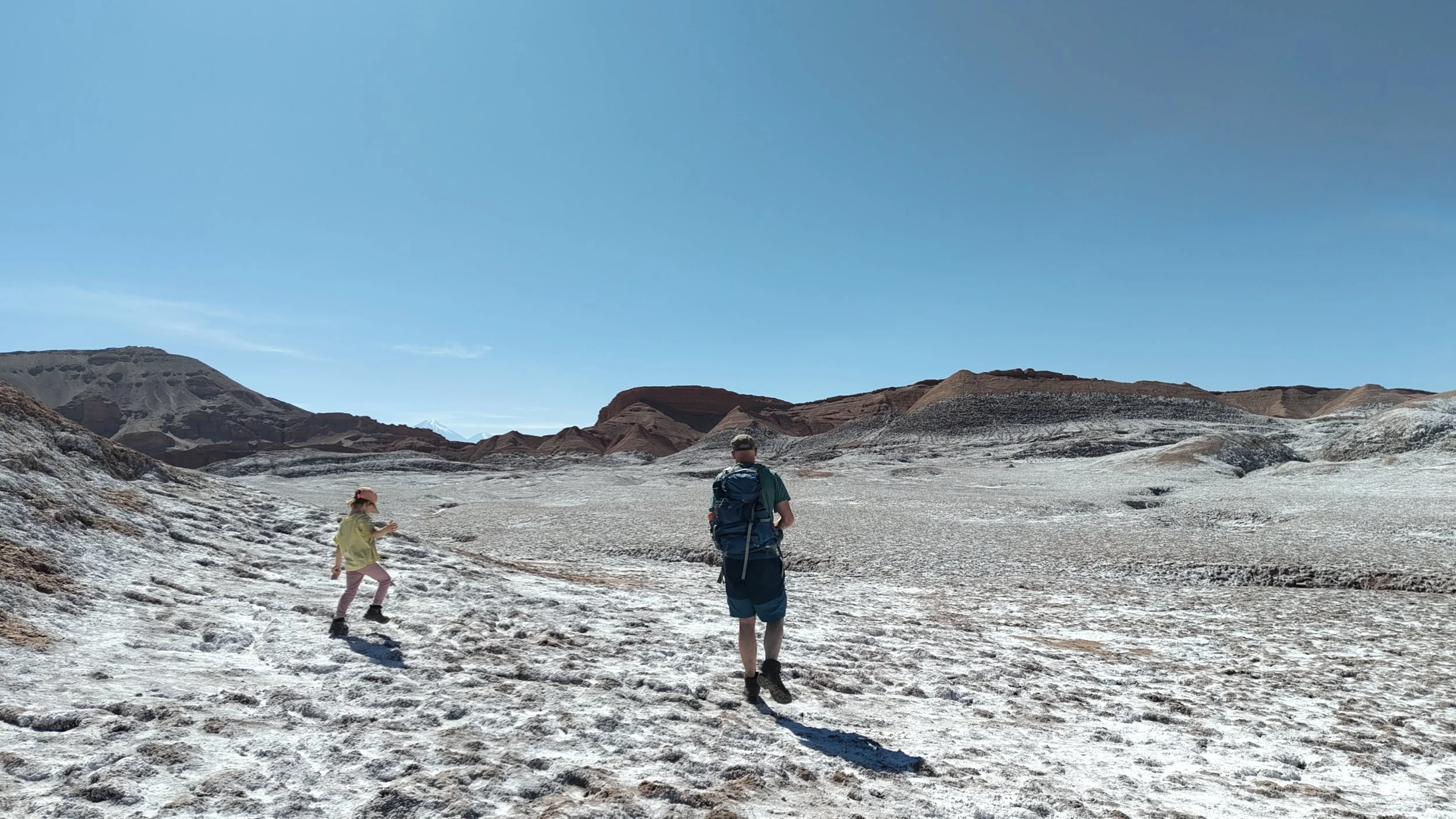 Zwei Erwachsene und ein Kind beim Wandern in einer kargen, trockenen Berglandschaft mit sandiger Oberfläche und farbigen Hügeln im Hintergrund, bei strahlendem Himmel.