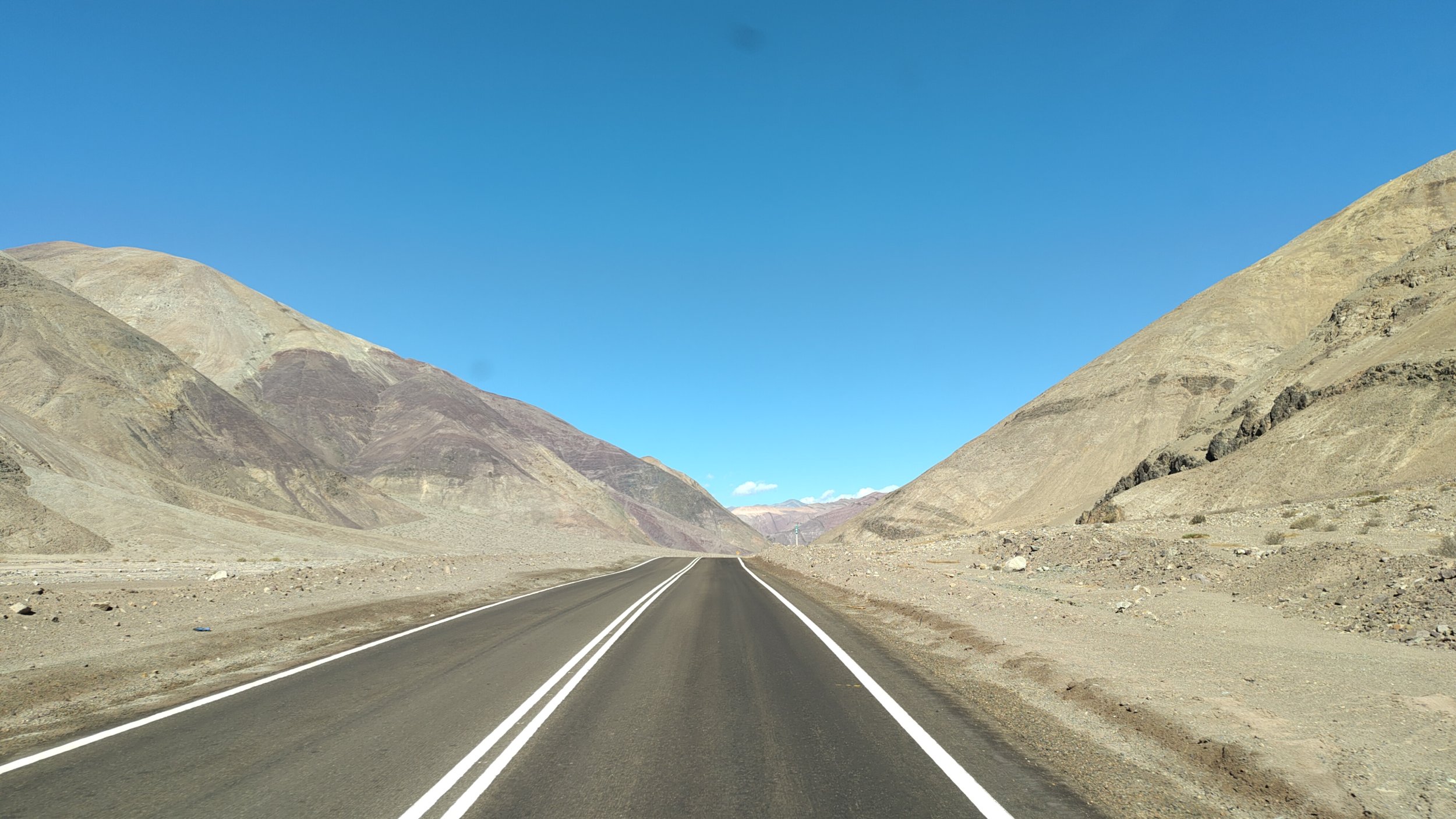 Eine stillgelegene Straße in einer kargen, bergigen Wüste mit zwei parallelen weißen Linien auf der dunklen Asphaltstraße, umgeben von trockenen, felsigen Hügeln unter klarem blauen Himmel.