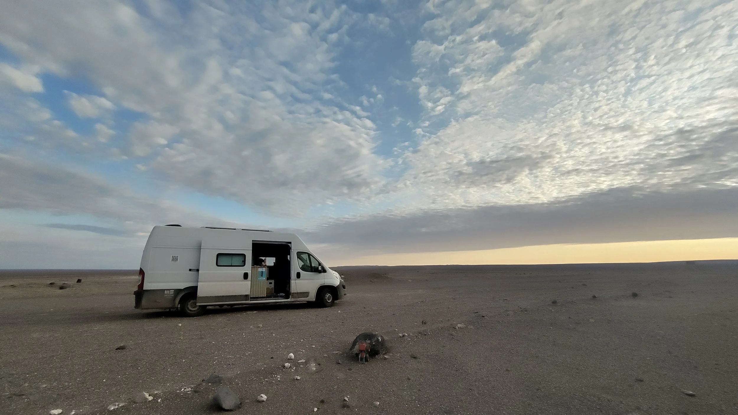 Ein weißer Van parkt auf einer kargen, staubigen Landschaft unter einem bewölkten Himmel, mit Blick auf die untergehende Sonne am Horizont.