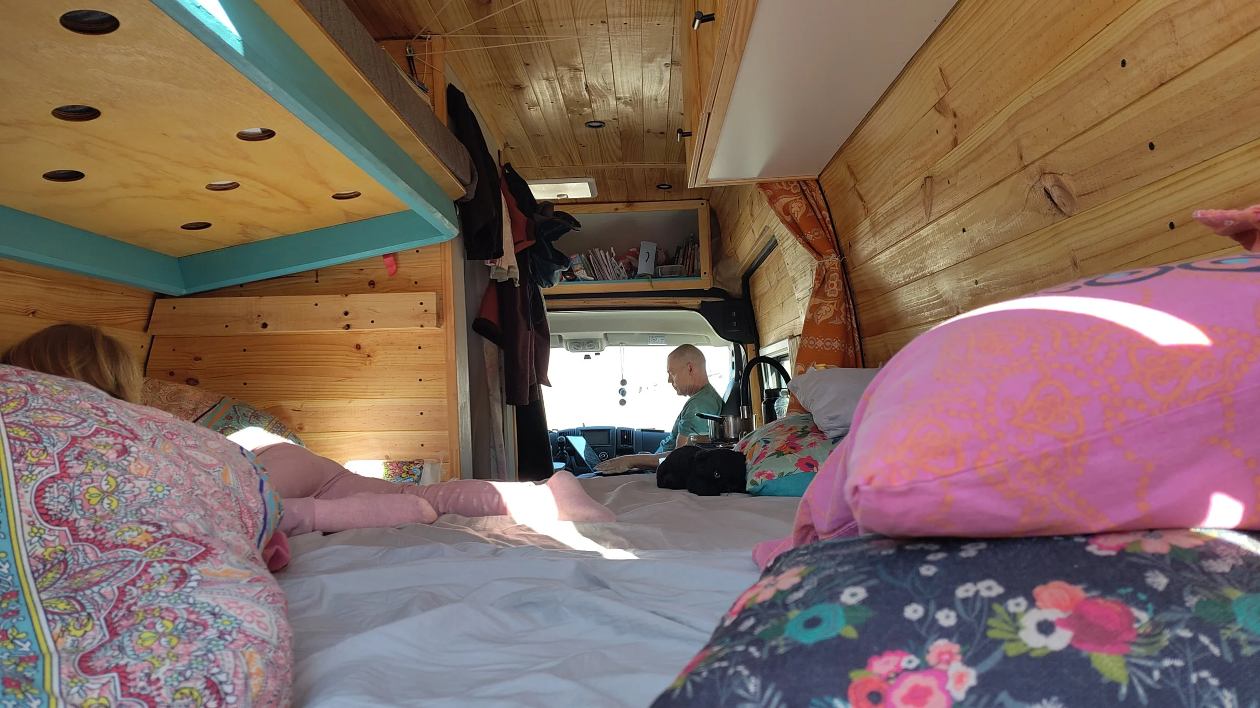 Innenansicht eines umgebauten Wohnmobils mit Holzinterieur, bettein Rückseite und Fahrer. Zwei Personen sind anwesend, eine sitzt auf einem Bett und eine andere am Steuer.