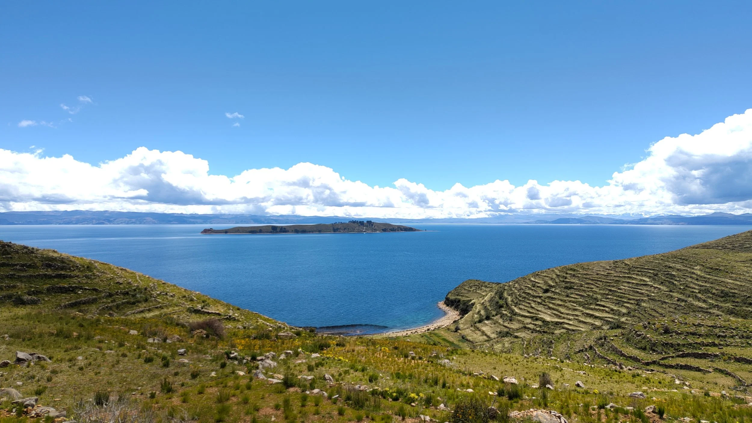 Blick auf einen großen See mit einer kleinen Insel im Wasser, umgeben von bewaldeten Hügeln und einer bewölkten blauen Himmelshintergrund.
