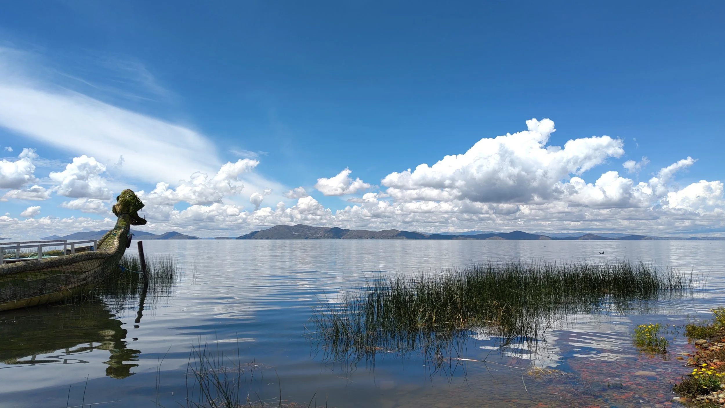 Blick auf einen See mit Bergen im Hintergrund, Wolken am Himmel, und eine Drachenfigur im Wasser im Vordergrund.