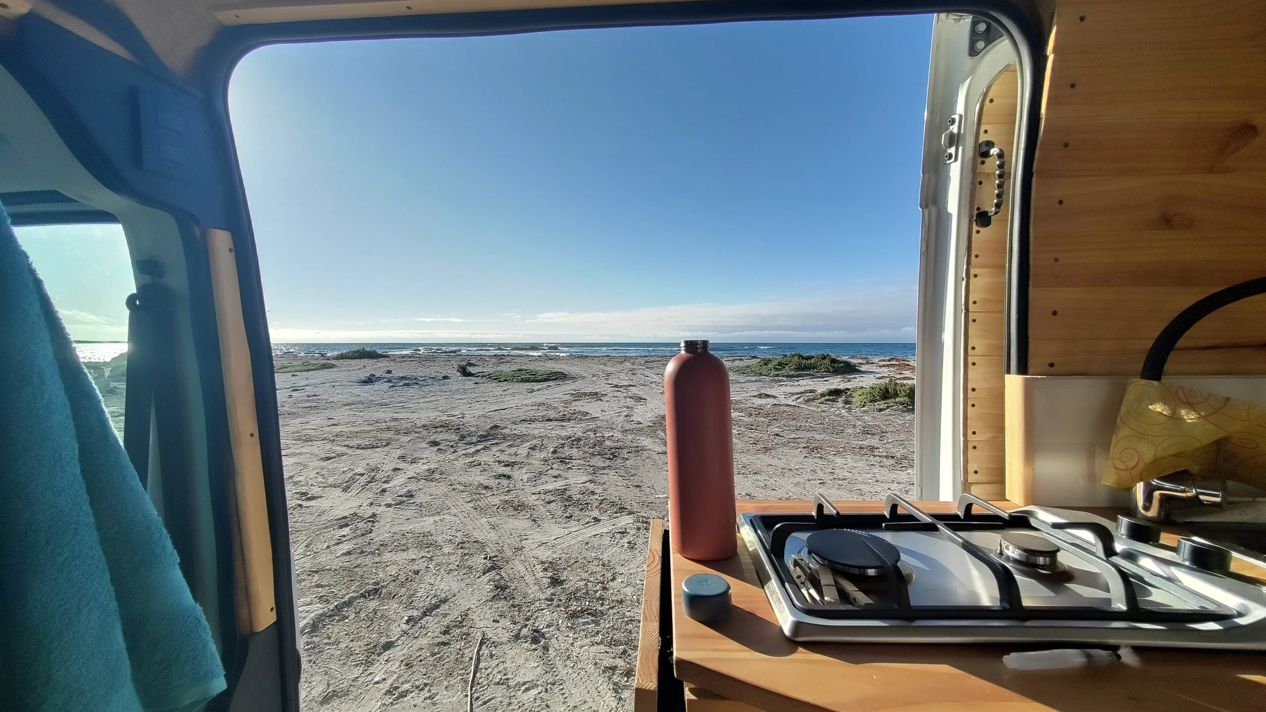 Blick aus dem Inneren eines Minivans mit Aussicht auf einen Strand und das Meer, Küche im Vordergrund, blauer Himmel.