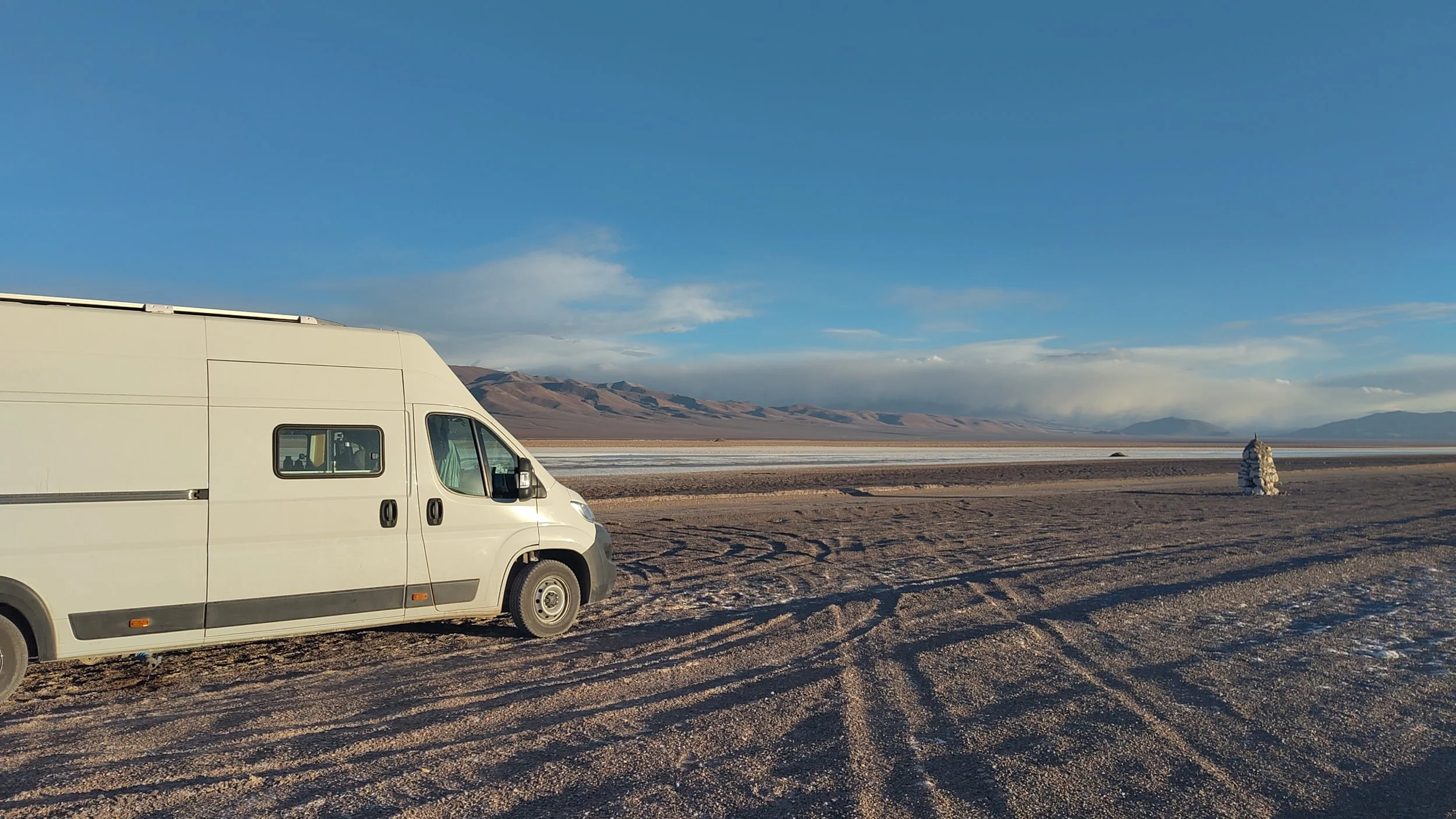 Ein weißer Campingwagen steht in einer Wüstenlandschaft mit Salzfläche, Berge im Hintergrund und einem blauen Himmel, während die Sonne scheint.
