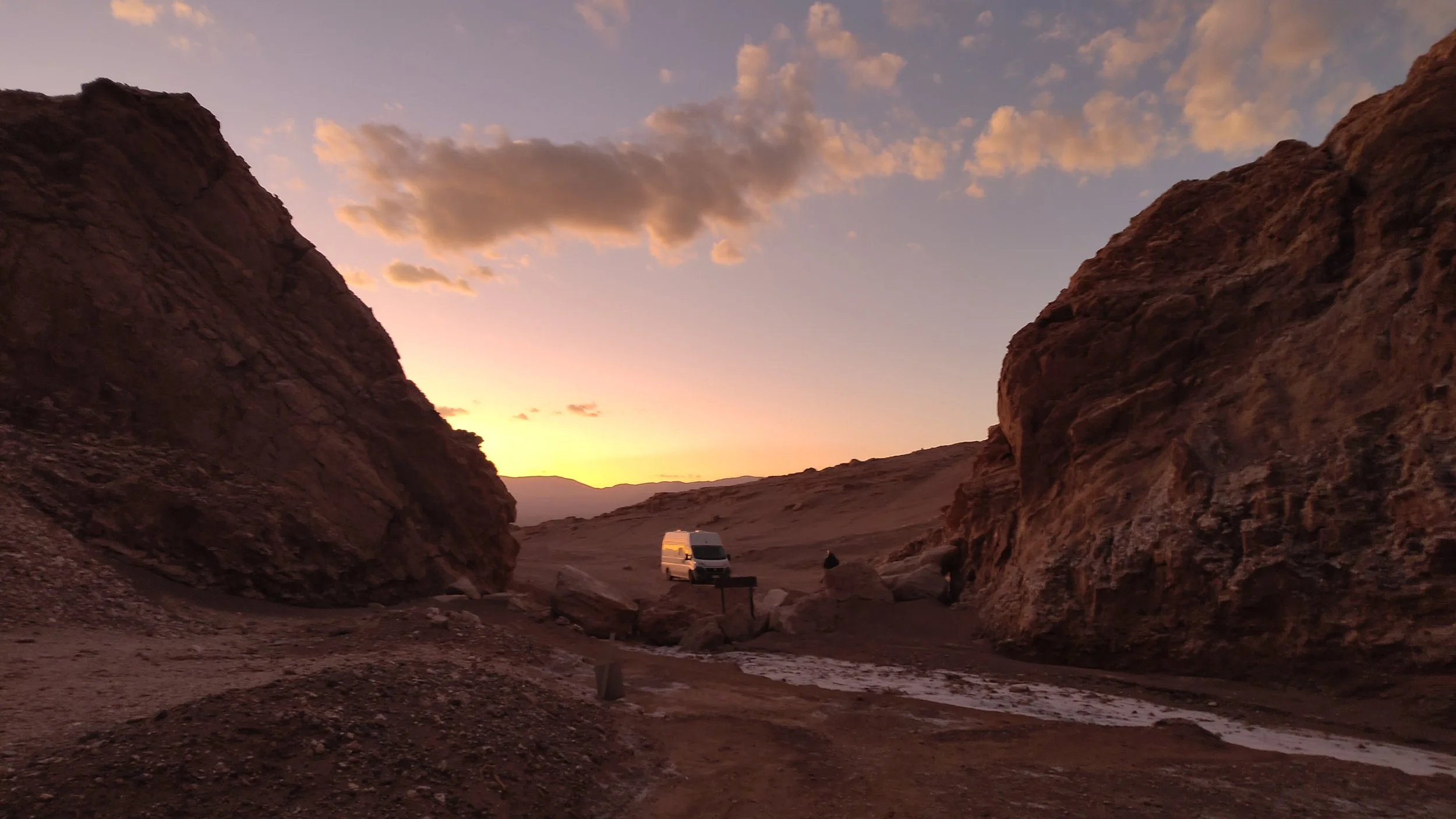 Wüste bei Sonnenuntergang mit Felsformationen und einem weißen Van im Hintergrund.