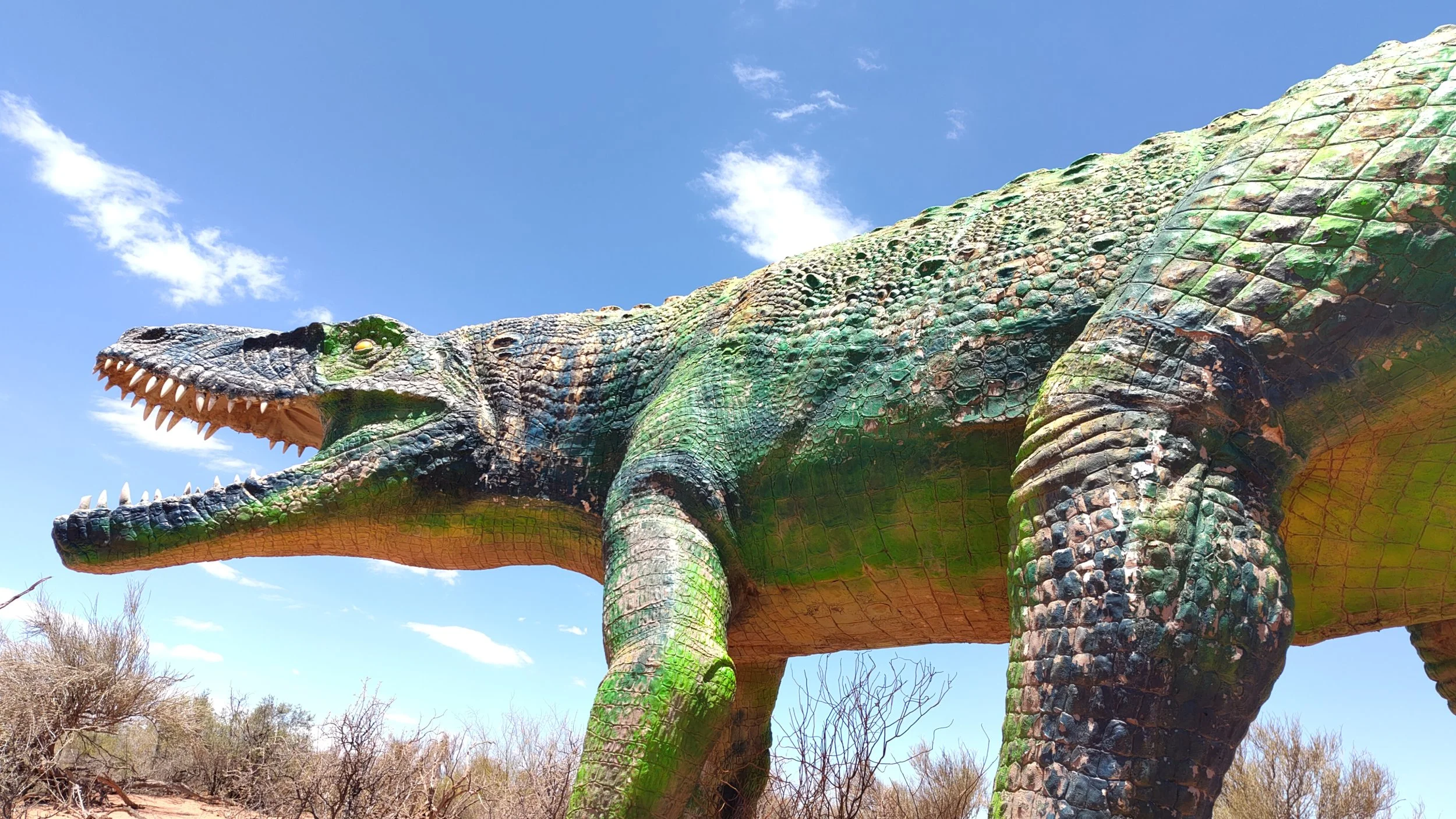 Aufblasbares Dinosauriermodell mit grüner und grauer Haut, offene Maul mit sichtbaren Zähnen, im Freien bei blauem Himmel.