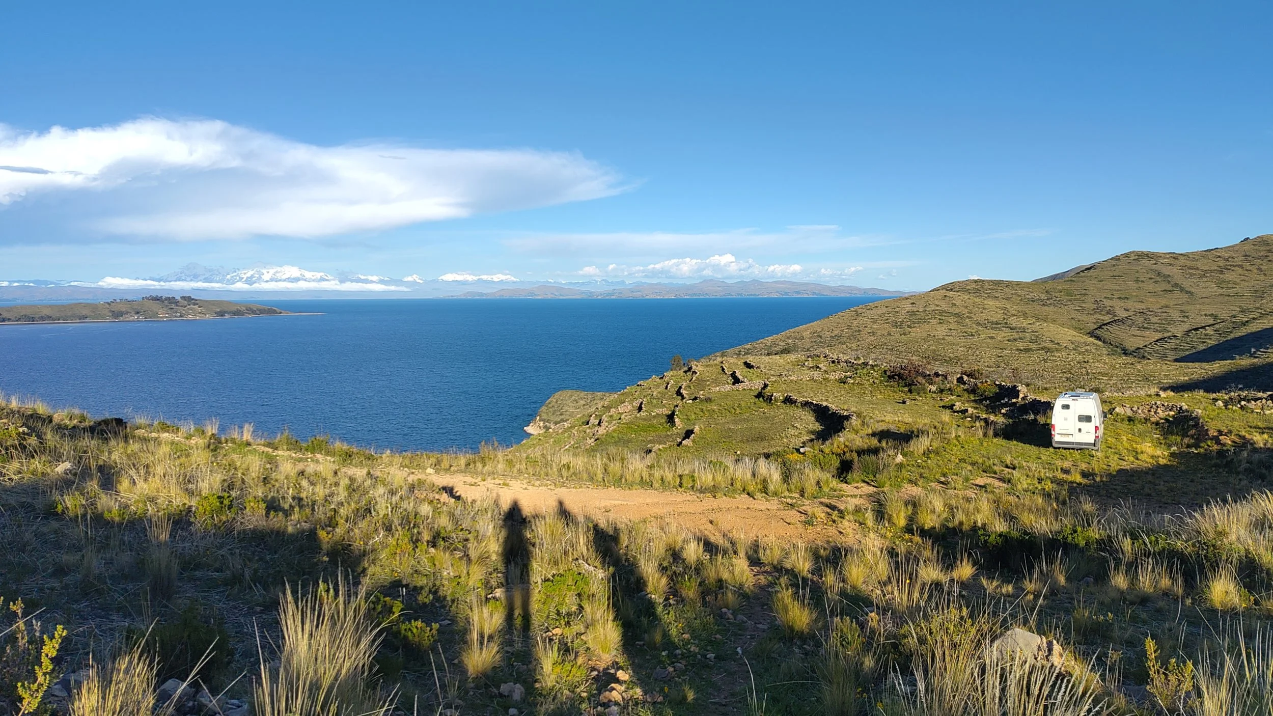 Blick auf einen großen See mit Bergen im Hintergrund, Gräser und Bus im Vordergrund.