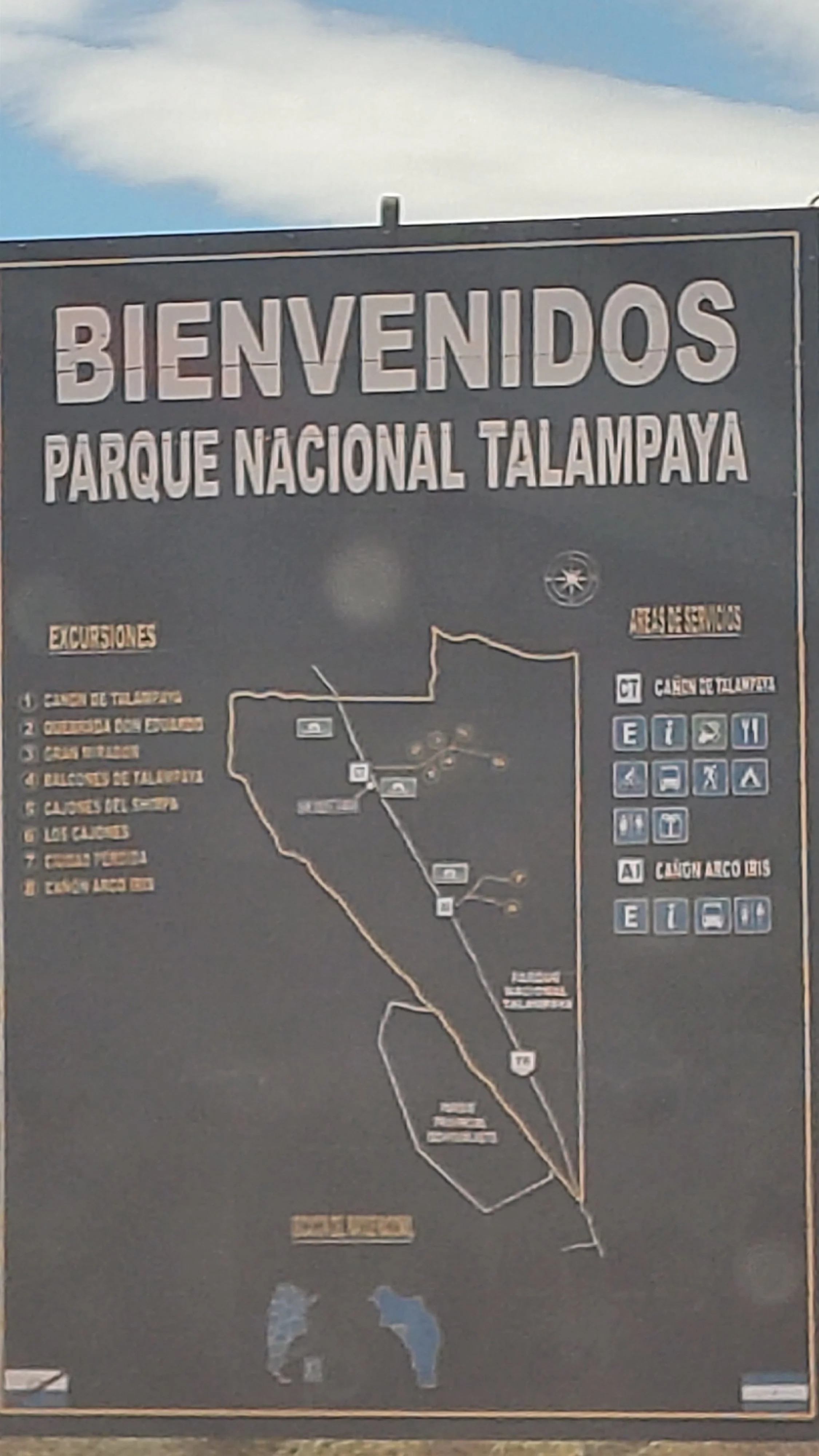 Hinweisschild des Nationalparks Talampaya mit einer Karte der verschiedenen Touren, Informationen, Symbole für Einrichtungen und eine Übersicht der Wege im Park, auf dem blauen Himmel zu sehen.