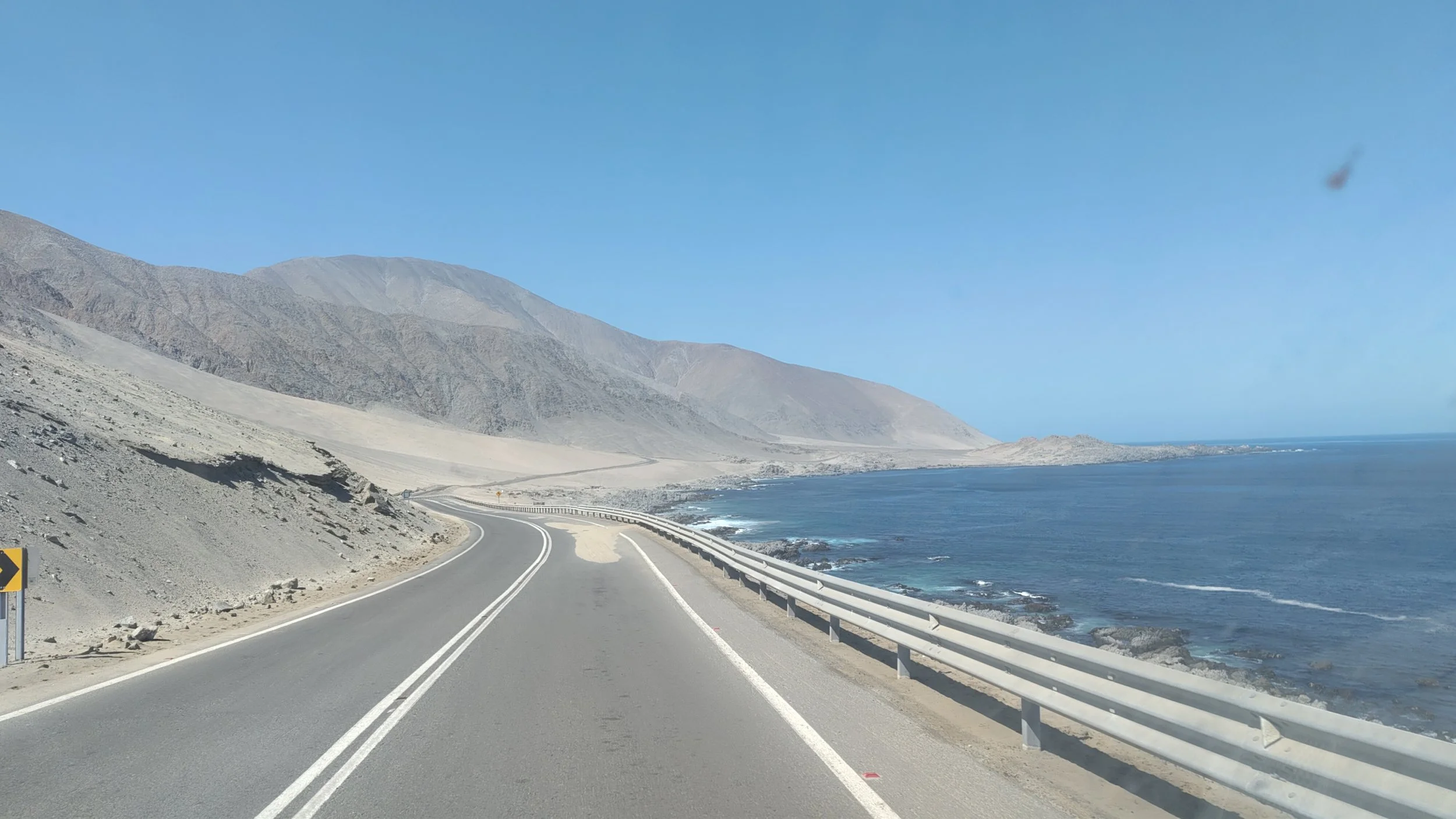 Straße entlang der Küste mit Bergen im Hintergrund und dem Meer auf der rechten Seite, sonniger Himmel.
