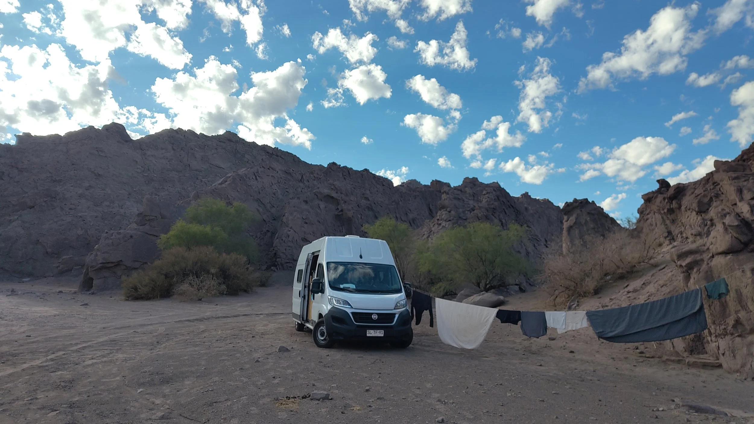 Ein Van in einer Wüstenlandschaft mit Felsen, Bäumen und Wäsche, die im Freien hängt, bei blauem Himmel mit Wolken.