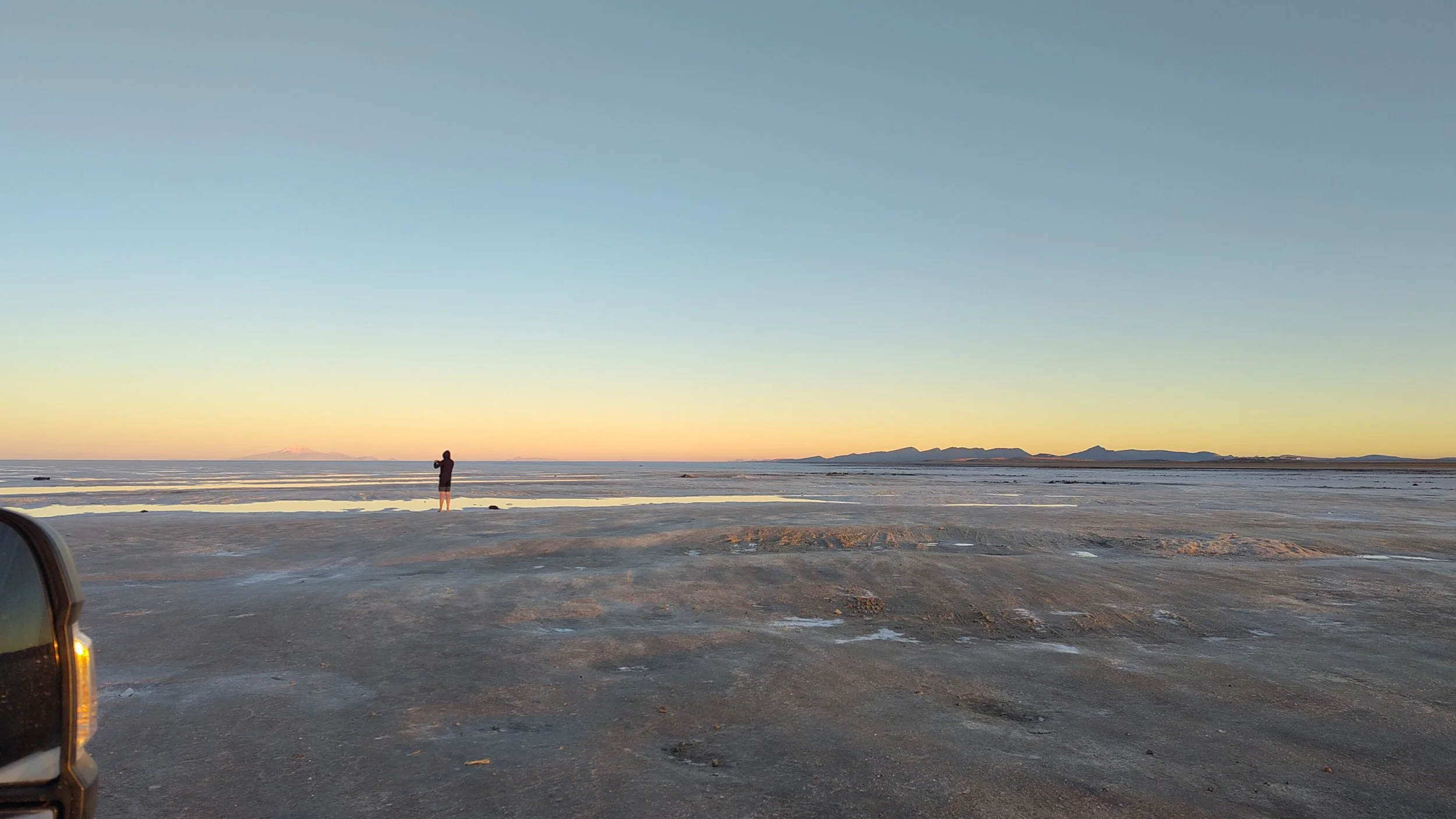Person steht am Ufer eines Sees bei Sonnenuntergang, im Hintergrund Berge, im Vordergrund eine flache, trockene Landschaft