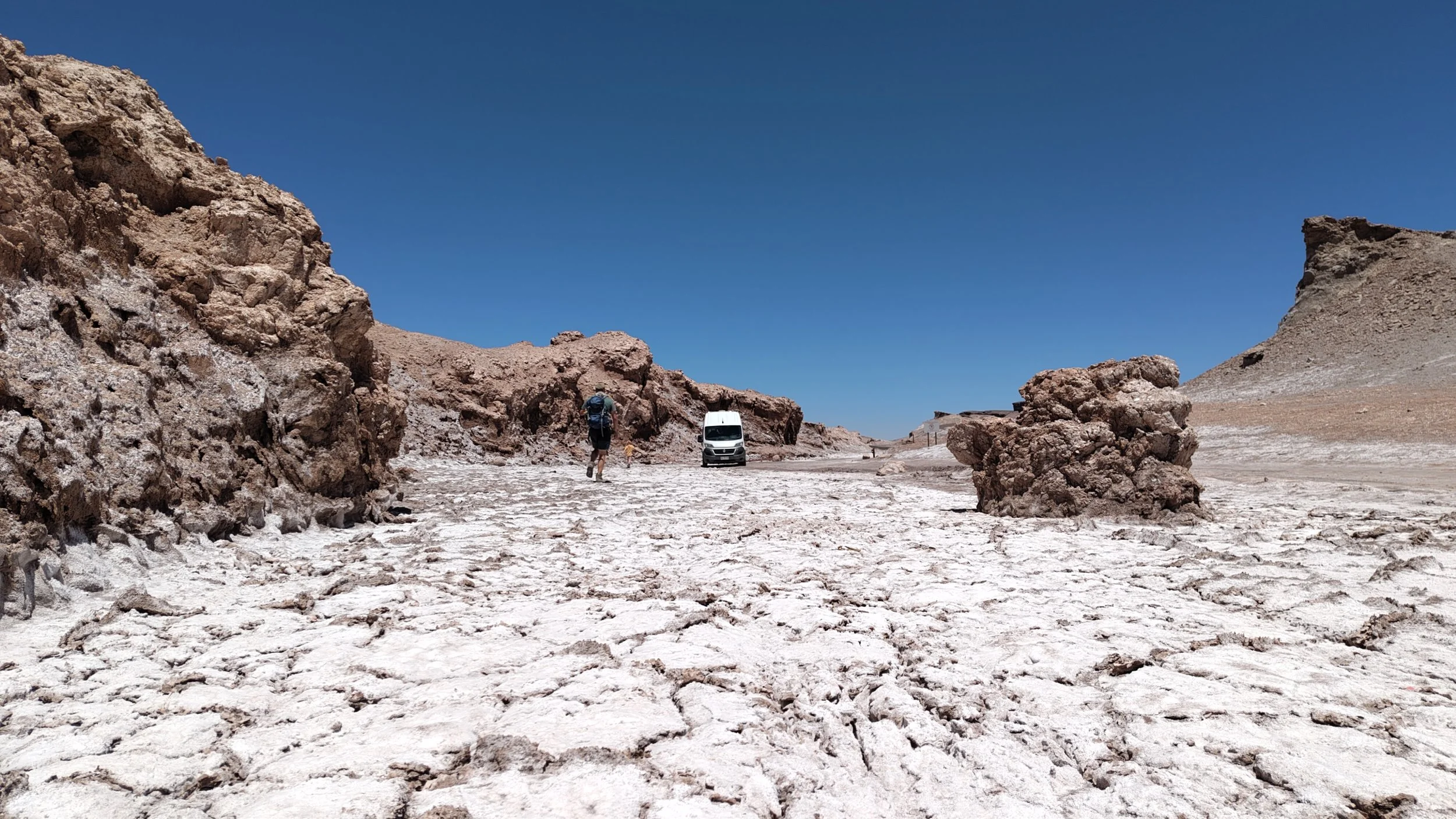 Ein Wanderer mit Rucksack geht durch eine trockene, felsige Landschaft mit großen Gesteinsformationen und klarem blauen Himmel.