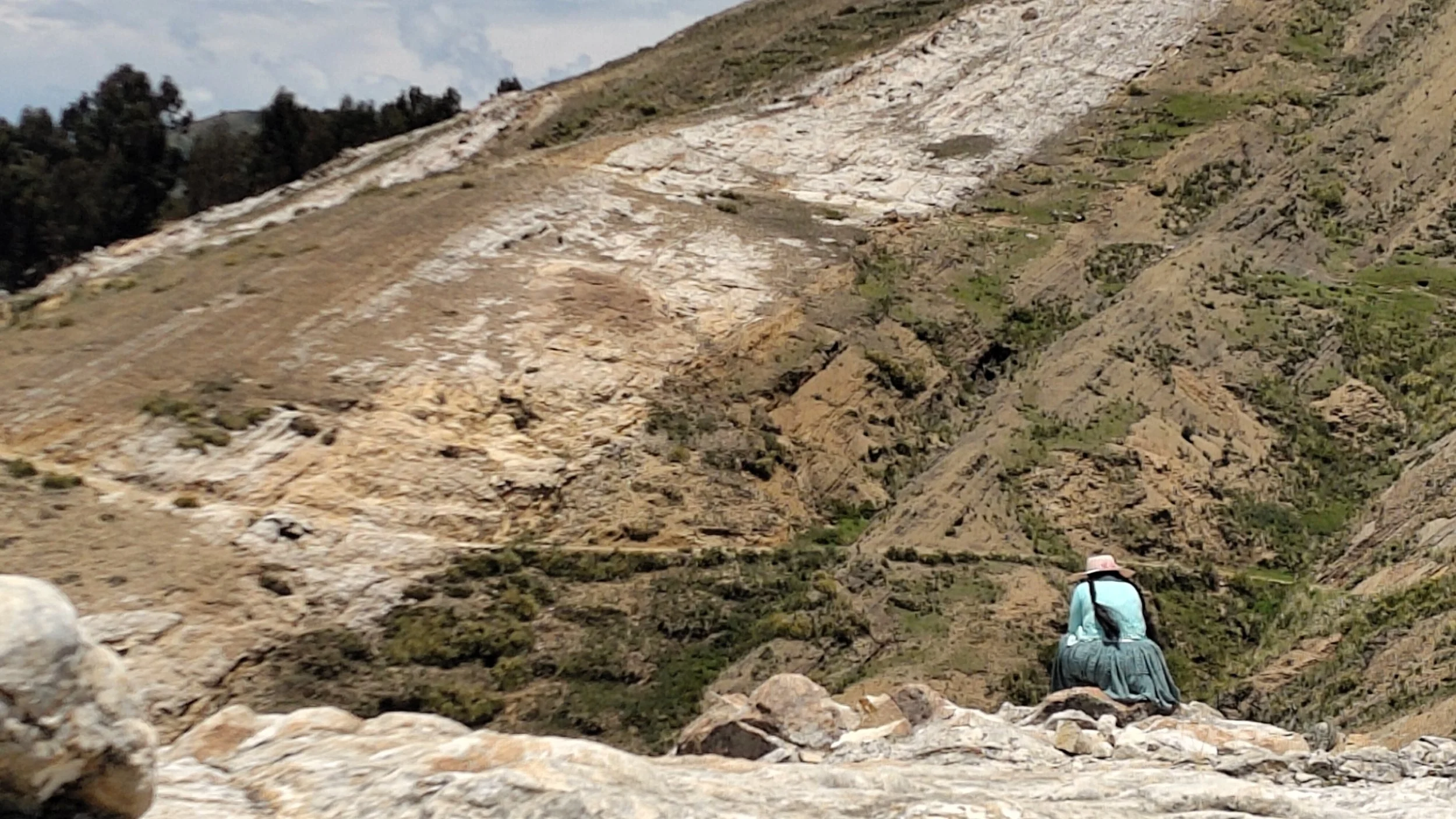 Ein Wanderer in einem blauen Hemd und einem Hut sitzt auf Steinen in einer felsigen Gebirgsregion, umgeben von steilen Berghängen und trockenem Gelände.
