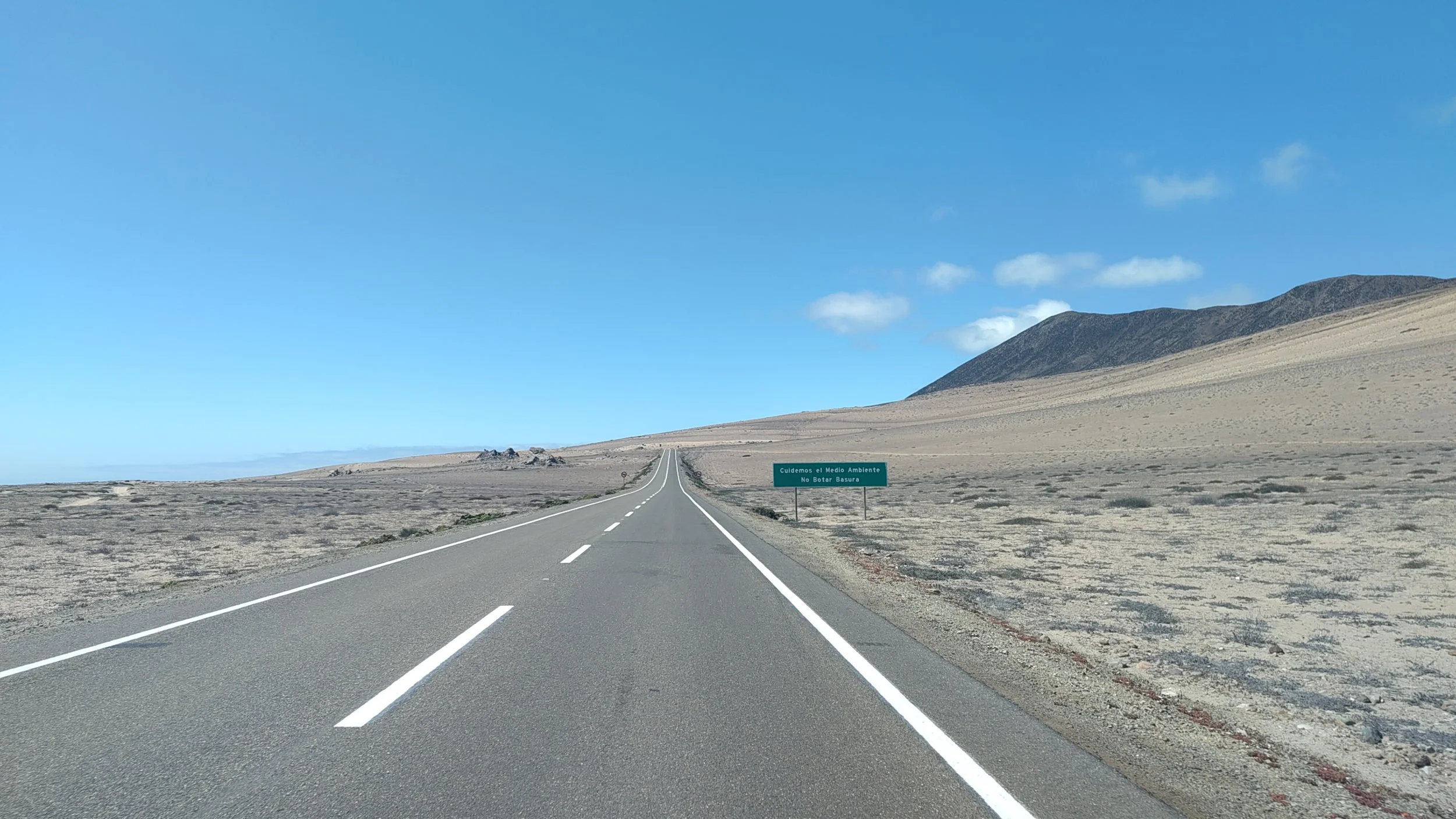 Leere Straße in einer Wüstenlandschaft mit Bergen im Hintergrund und einem grünen Schild, das auf Spanisch steht.