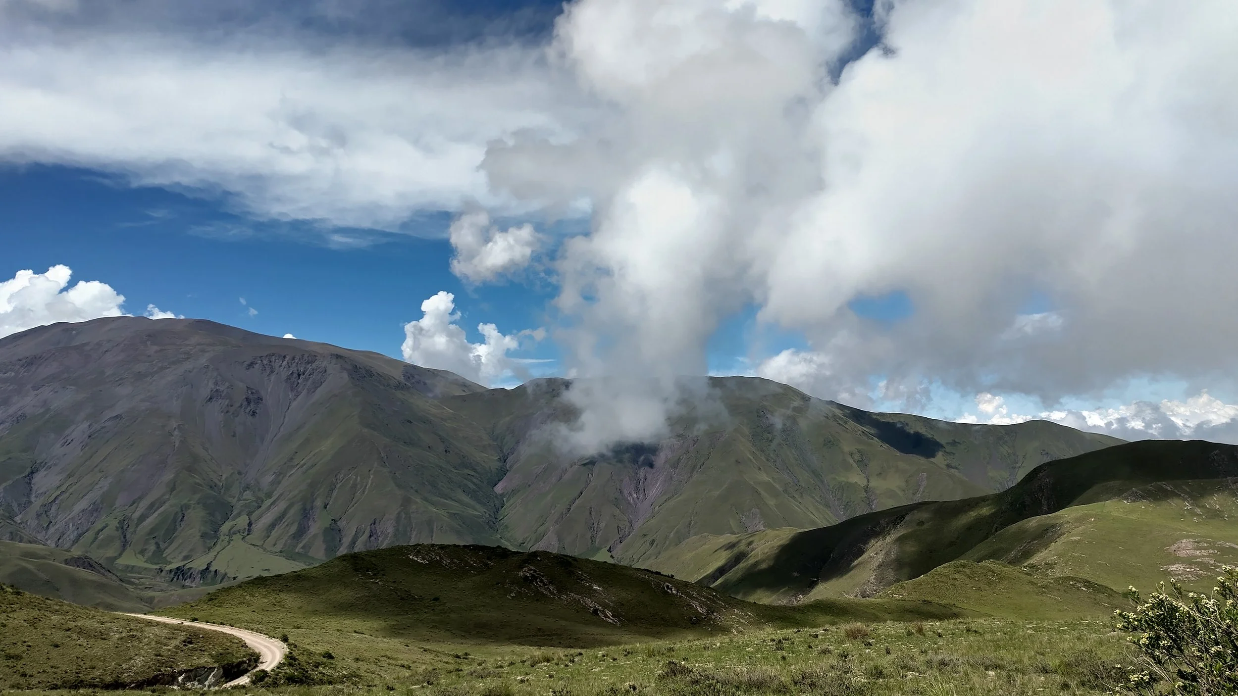 Grüne Hügel mit einer Rodelstraße, im Hintergrund Berge mit Wolken im Himmel.