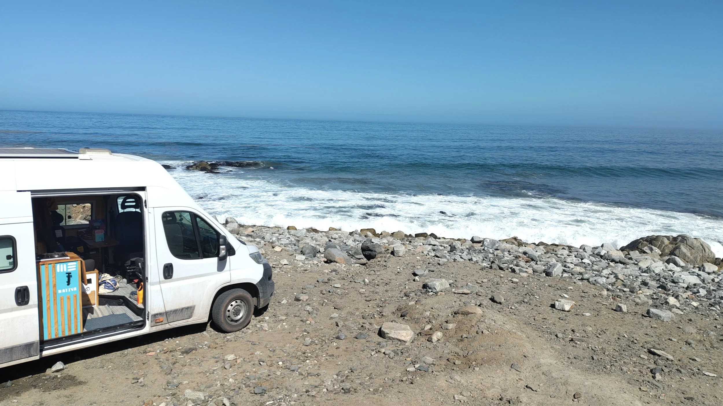 Weißer Van am Strand mit offenem Seitentür, darin sind Möbel und Gegenstände sichtbar, während im Hintergrund das Meer bei sonnigem Himmel zu sehen ist.