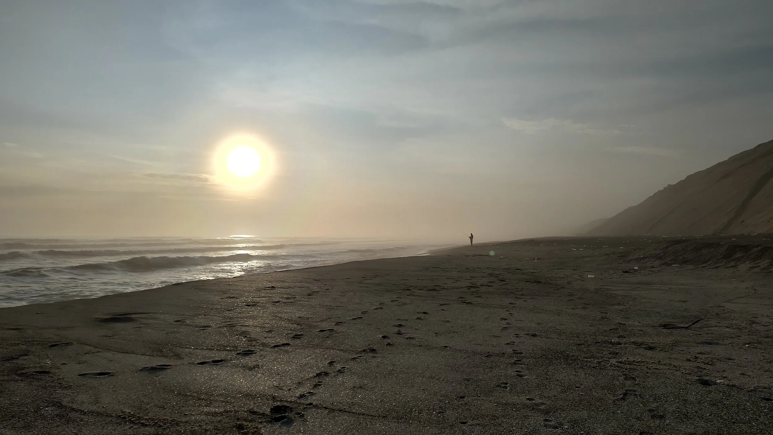 Ein einsamer Angler steht am Strand bei Sonnenuntergang, das Meer ist ruhig und die Küste ist von Klippen umgeben.