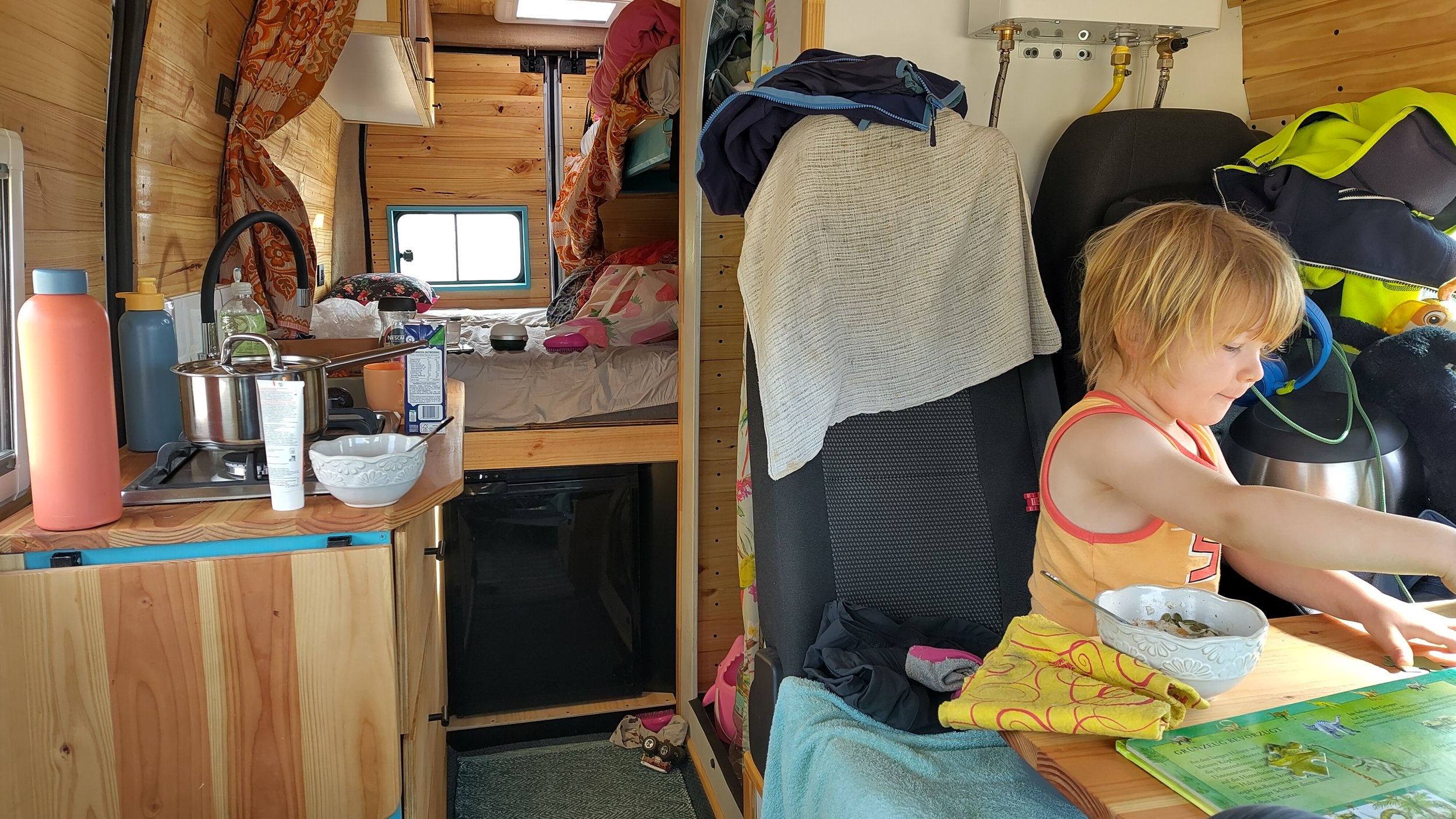 Innenansicht eines kleinen Holzhauses oder Wohnwagens mit einem Schlafraum im Hintergrund, einer Küchenzeile im Vordergrund, eine kleine Frau beim Essen am Tisch, und viele persönliche Gegenstände, Kleidung und Haushaltsartikel im Raum verteilt.
