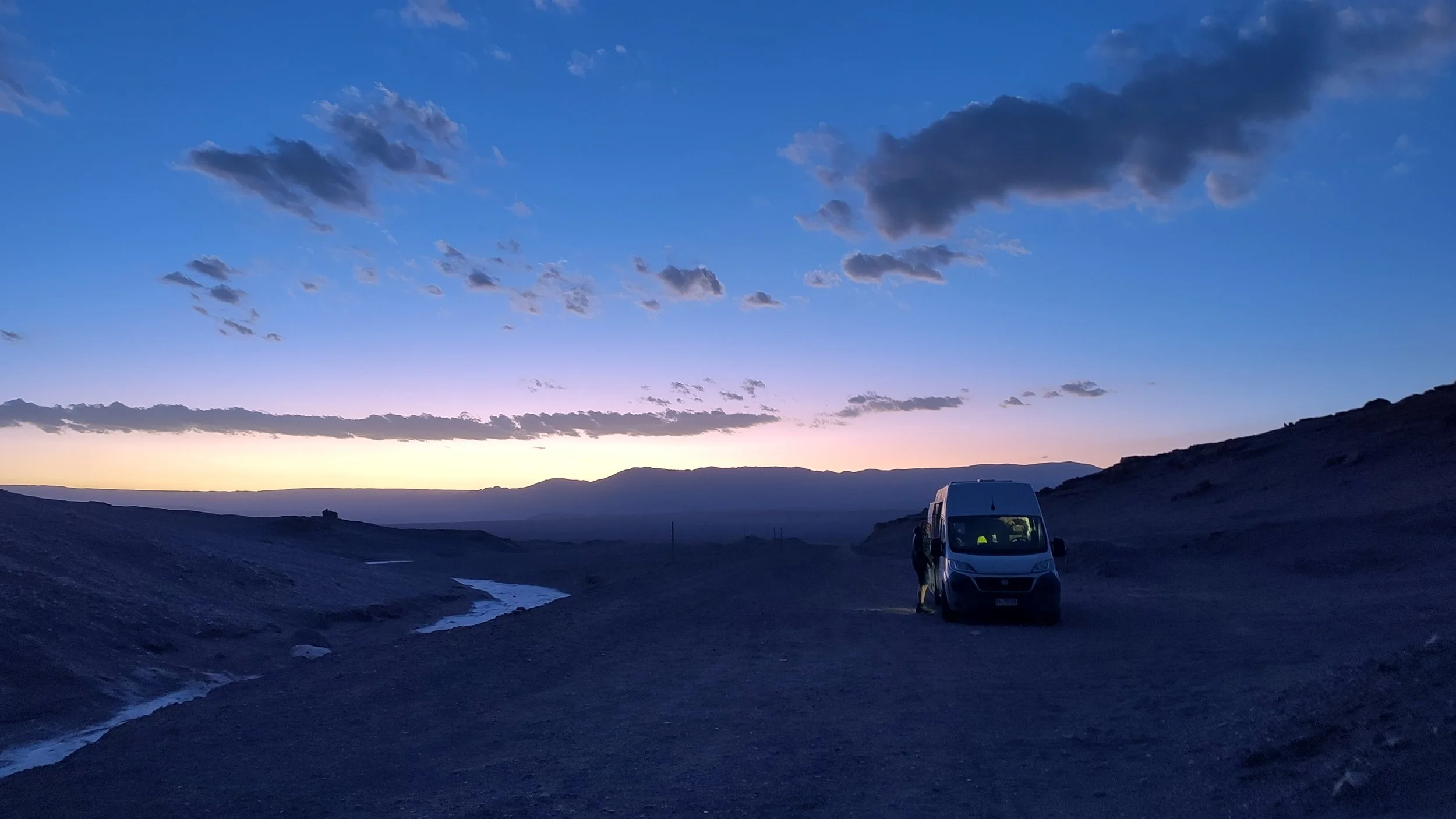 Ein Van steht auf einer ruhigen, offenen Landschaft bei Sonnenuntergang; Himmel ist blau mit dunklen Wolken, Berge sind im Hintergrund, einige dünne Wasserläufe fließen durch die Landschaft, und eine Person steht neben dem Van im Schatten.