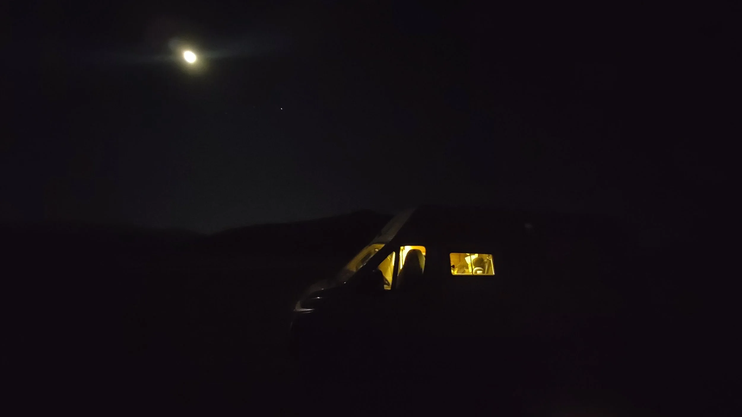 Ein Zelt in der Dunkelheit, beleuchtet durch eine warme Lichtquelle, mit einem hell erleuchteten Auto im Vordergrund und einem nächtlichen Himmel mit Mond und einem Stern darüber.