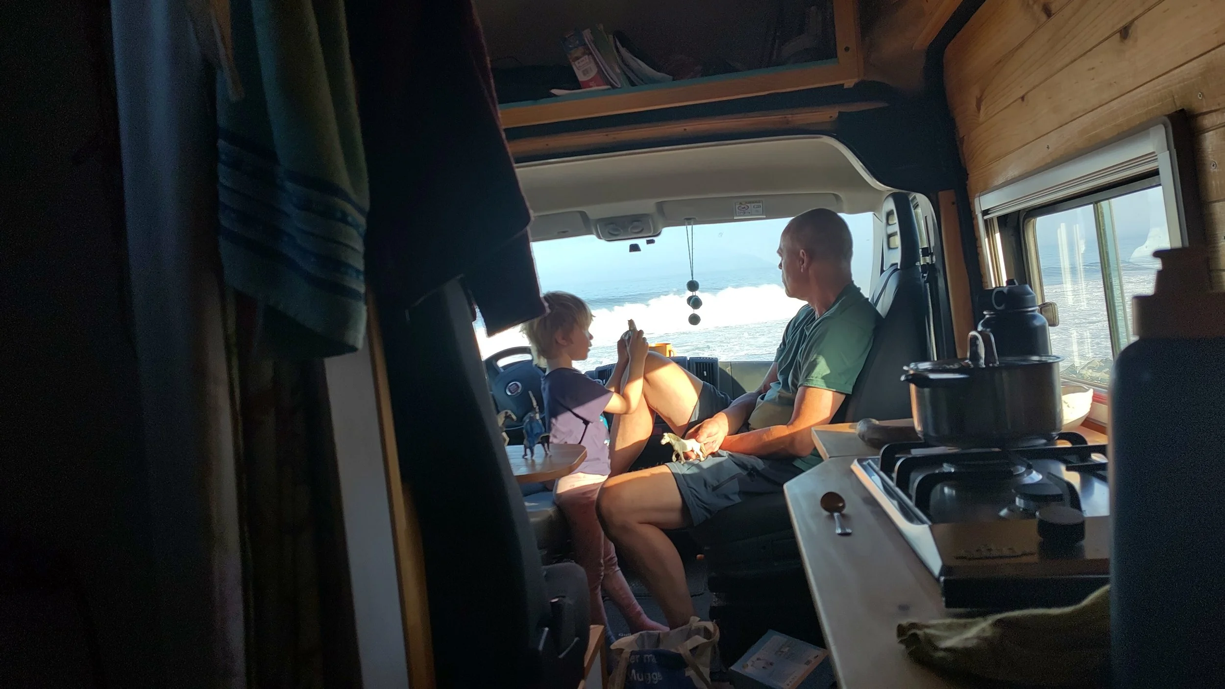 Ein Mann und ein Kind sitzen im Inneren eines Wohnmobils, während sie auf dem Wasser unterwegs sind, sichtbar durch die Frontscheibe, die Meer und Himmel zeigt.