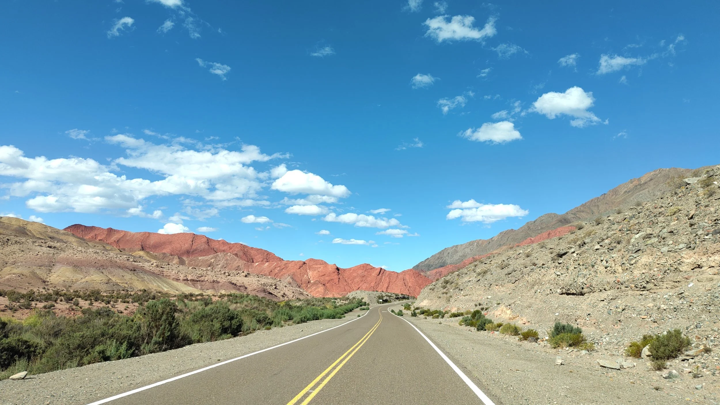 Eine zweispurige Straße verläuft durch eine wüstenartige Gebirgslandschaft mit roten und grauen Bergen unter einem blauen Himmel mit weißen Wolken.
