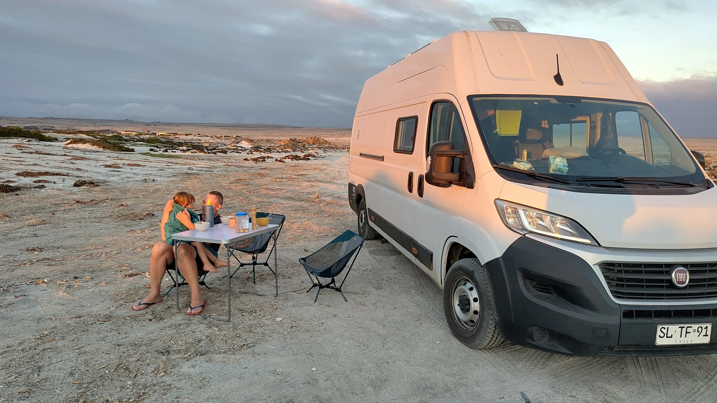Ein Wohnmobil parkt in einer Wüstenlandschaft bei Sonnenuntergang, während zwei Personen an einem kleinen Tisch essen und sich umarmen.