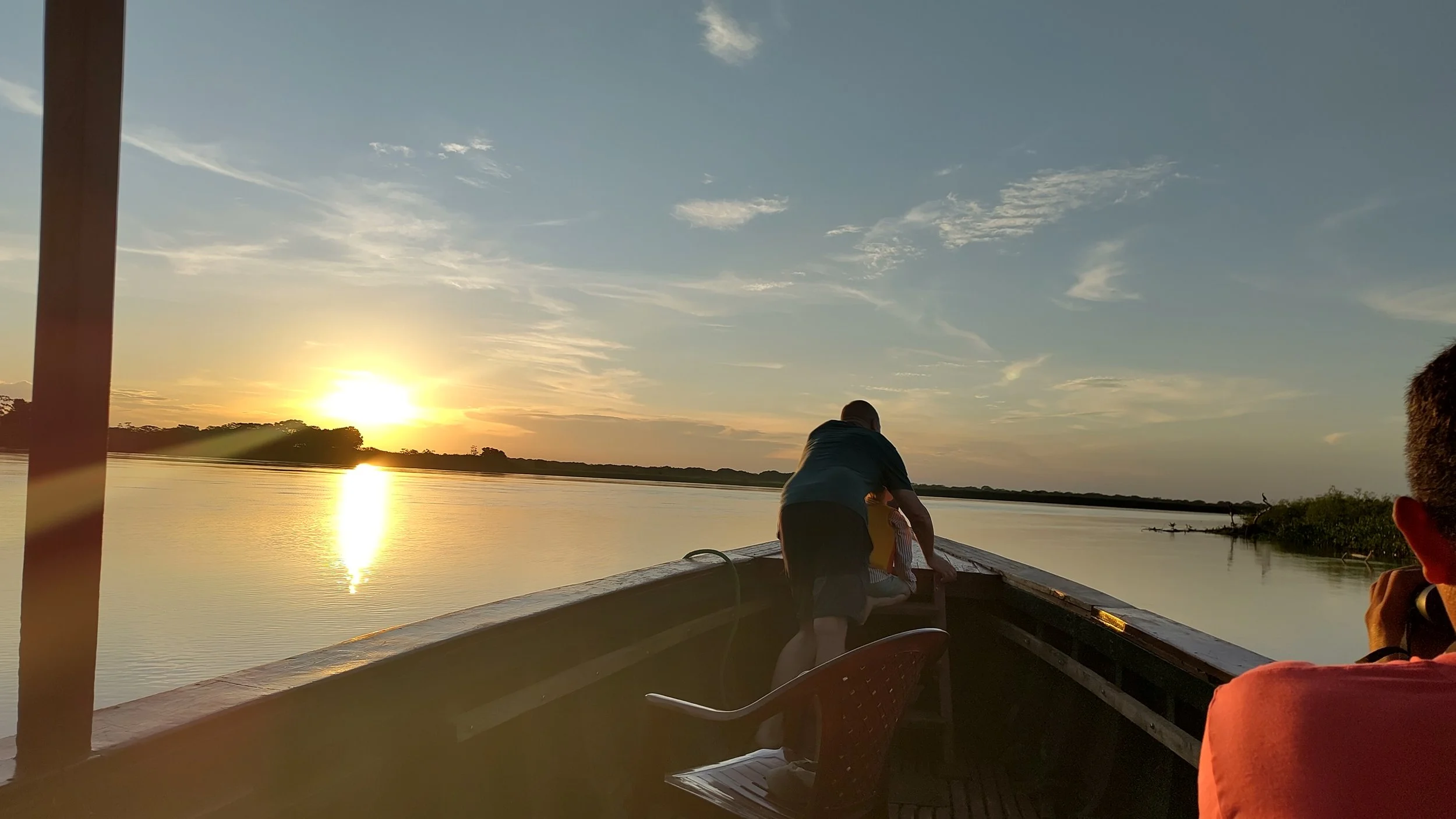 Menschen auf einem Boot bei Sonnenuntergang auf einem Fluss, einige sitzen und einer steht, die Landschaft ist ruhig mit wenigen Wolken am Himmel.