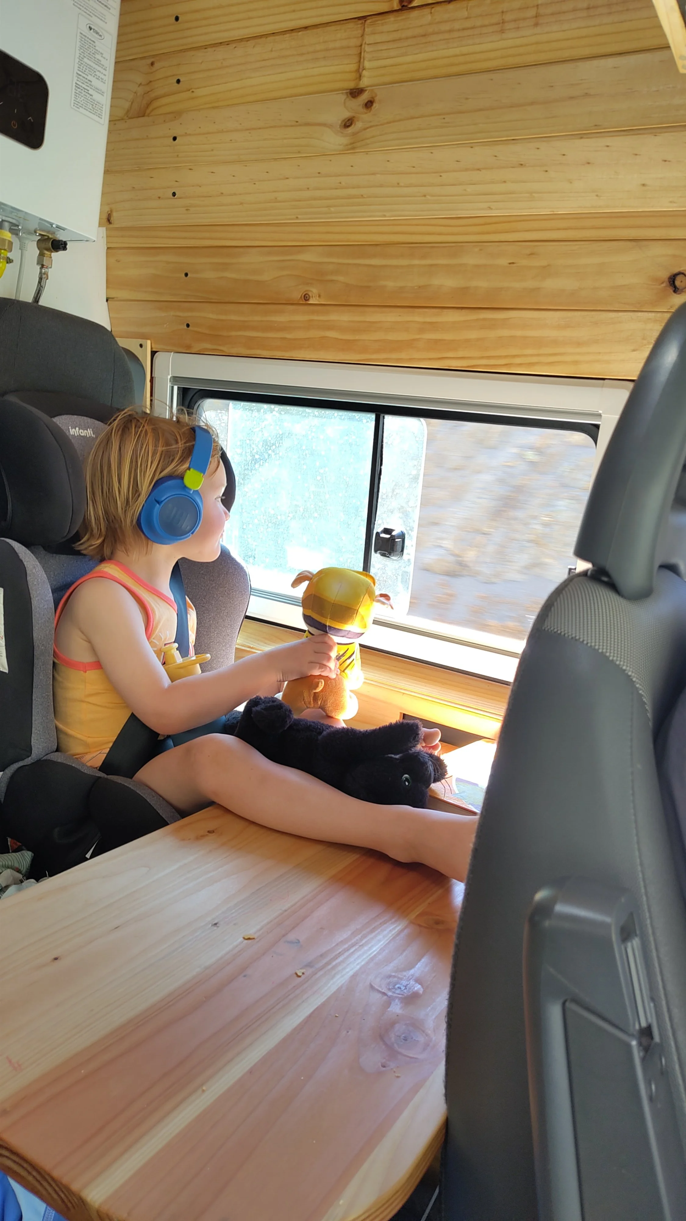 Ein kleines Mädchen mit blonden Haaren trägt Kopfhörer und sitzt in einem Kindersitz auf einem holzfarbenen Tisch. Es hält ein Stofftier in der Hand und schaut aus dem Fenster. Im Hintergrund sind Holzpaneelen an der Wand sichtbar.