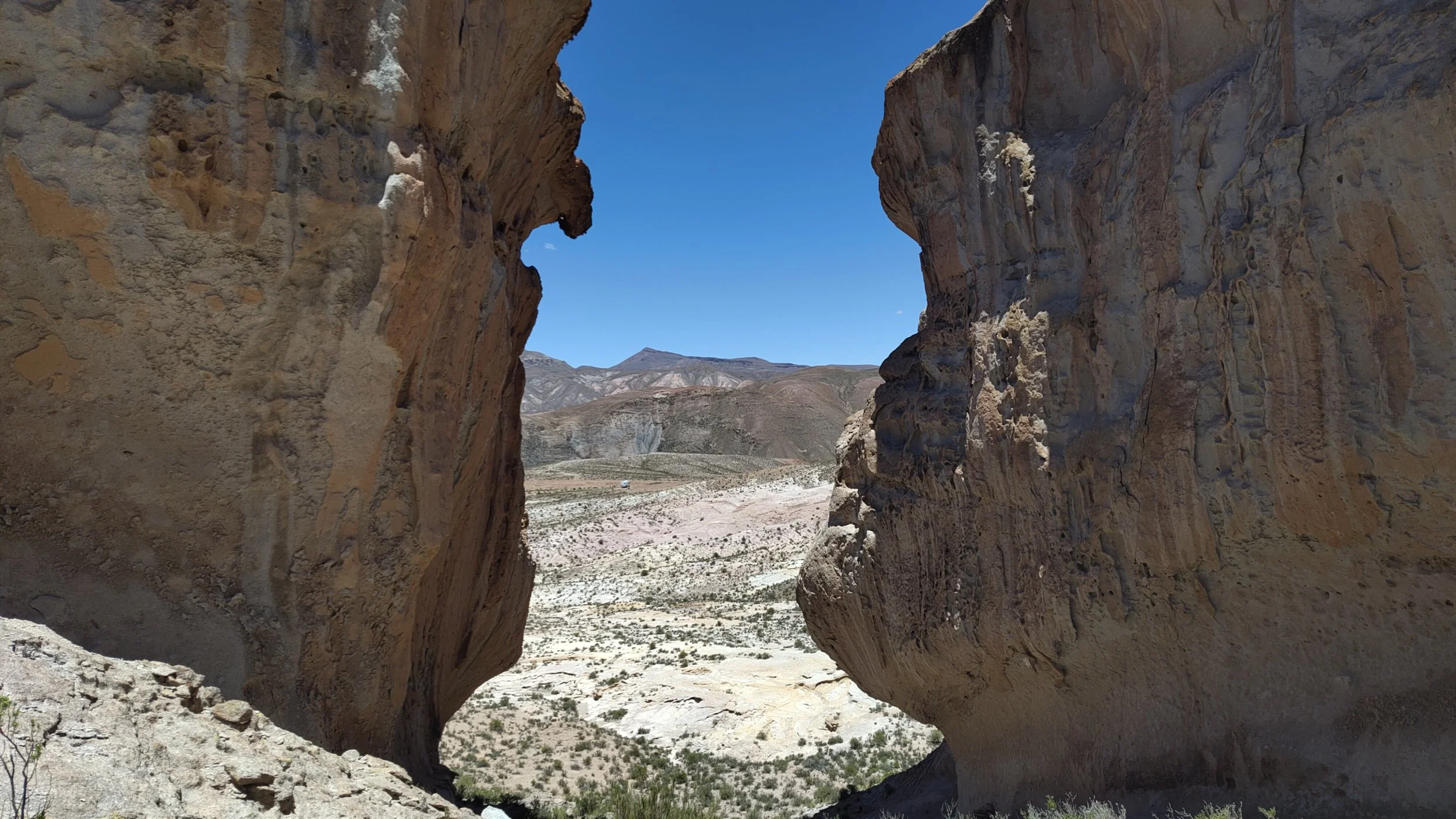 Felsen mit Durchblick in einer wüstenähnlichen Landschaft, Berge im Hintergrund, blauer Himmel.