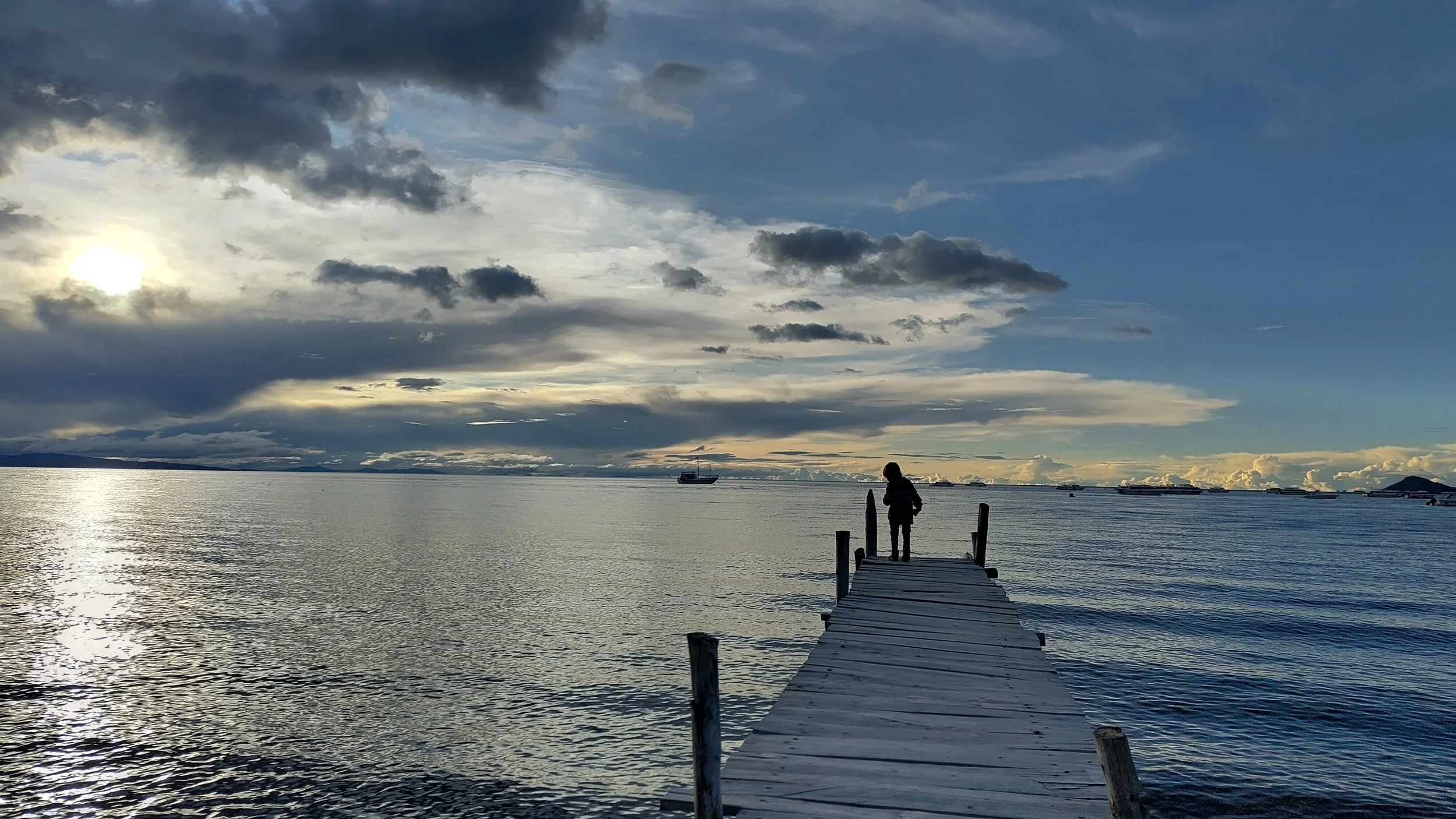 Person steht auf einem Steg am Wasser bei Sonnenuntergang, im Hintergrund sind Schiffe und Wolken am Himmel.