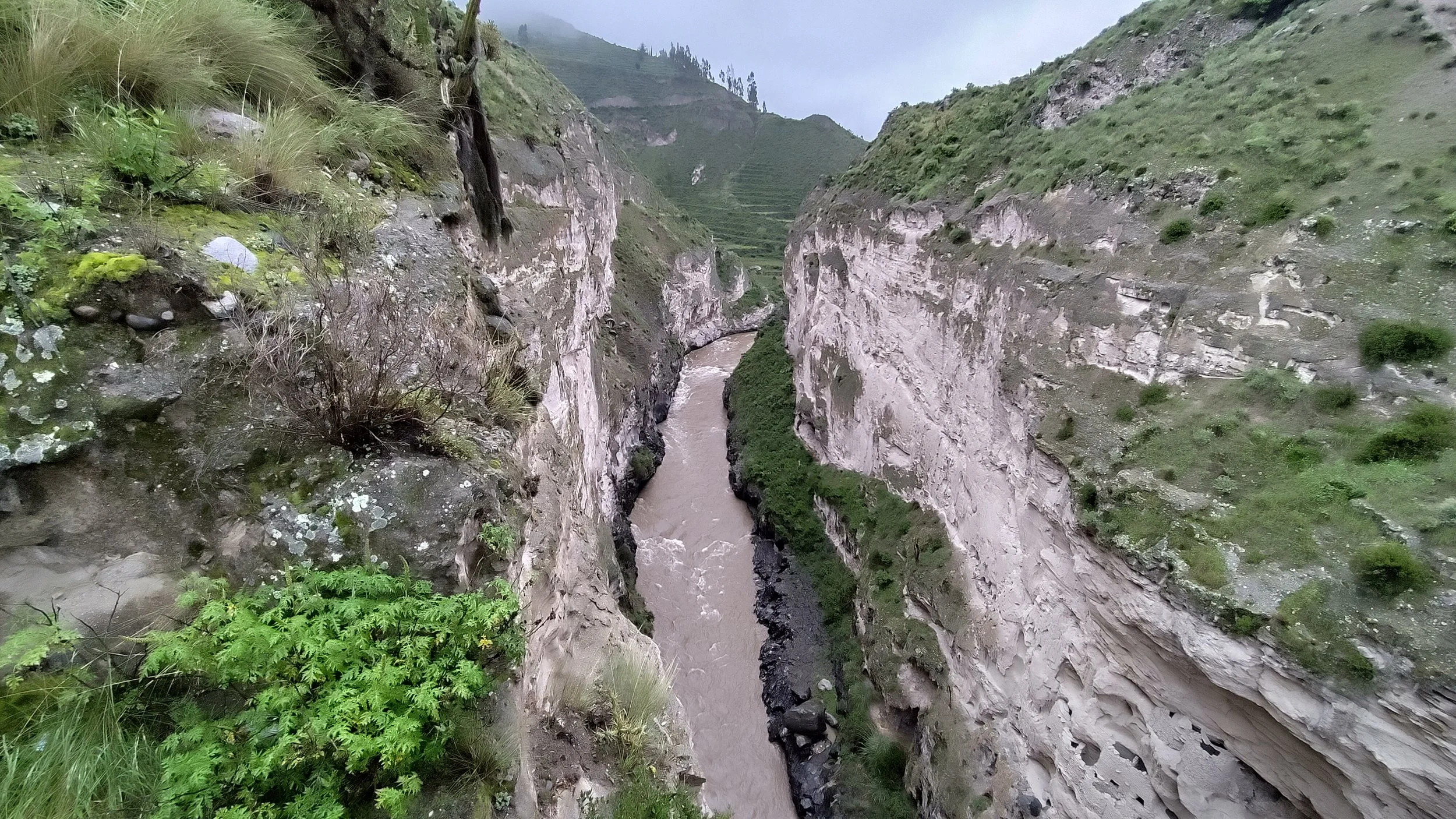 Eine enge, schroffe Schlucht mit einem fließenden Fluss in der Mitte, umgeben von grünen, bewaldeten Hängen und steilen Felswänden, bei bewölktem Himmel.