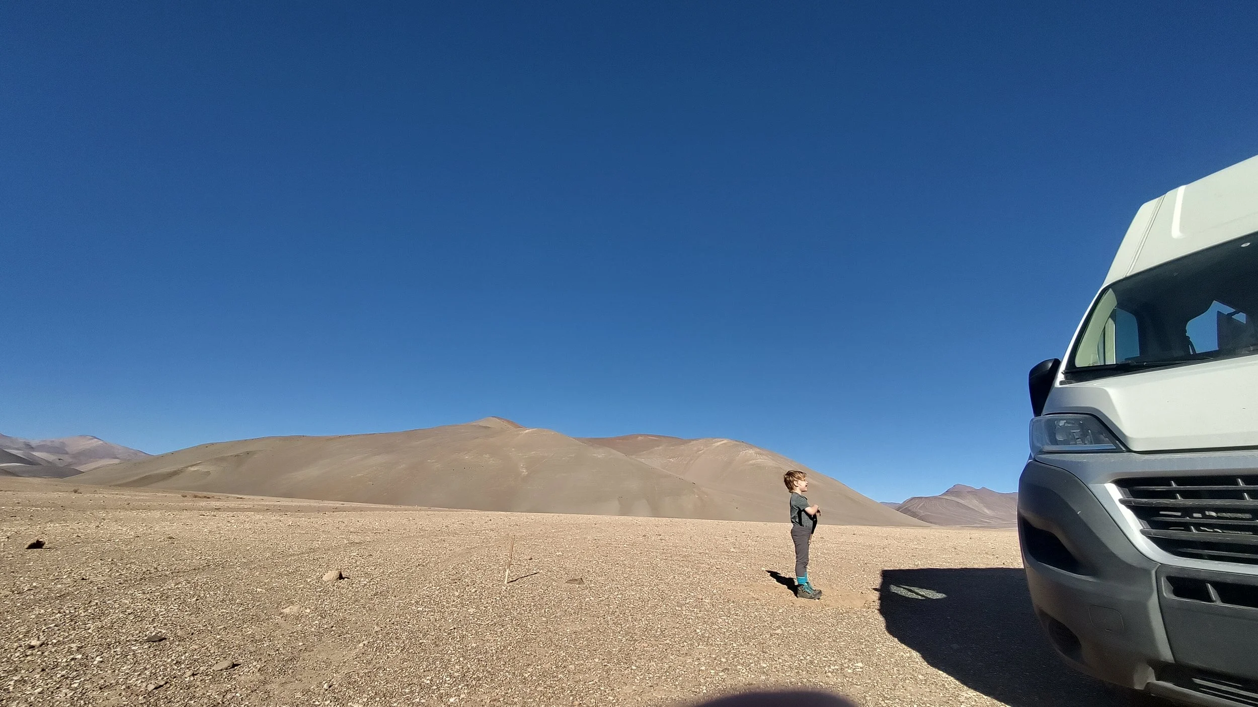 Ein junge steht in einer Wüste, im Hintergrund Berge und ein klarer blauer Himmel; auf der rechten Seite ist ein großes White-Kombi-Fahrzeug zu sehen.