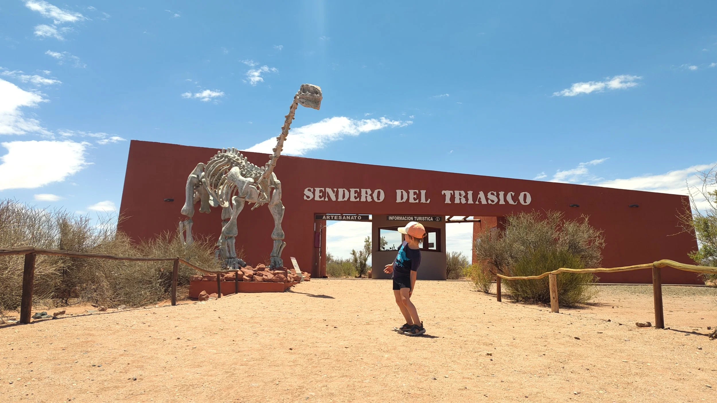 Ein Eingangsschild zum Dinosaurierpfad mit einem Dinosaurierskelett am Eingang, eine junge Person mit Hut steht davor, die Umgebung ist wüstenartig mit sandigem Boden und Sträuchern, der Himmel ist überwiegend klar mit einigen Wolken.