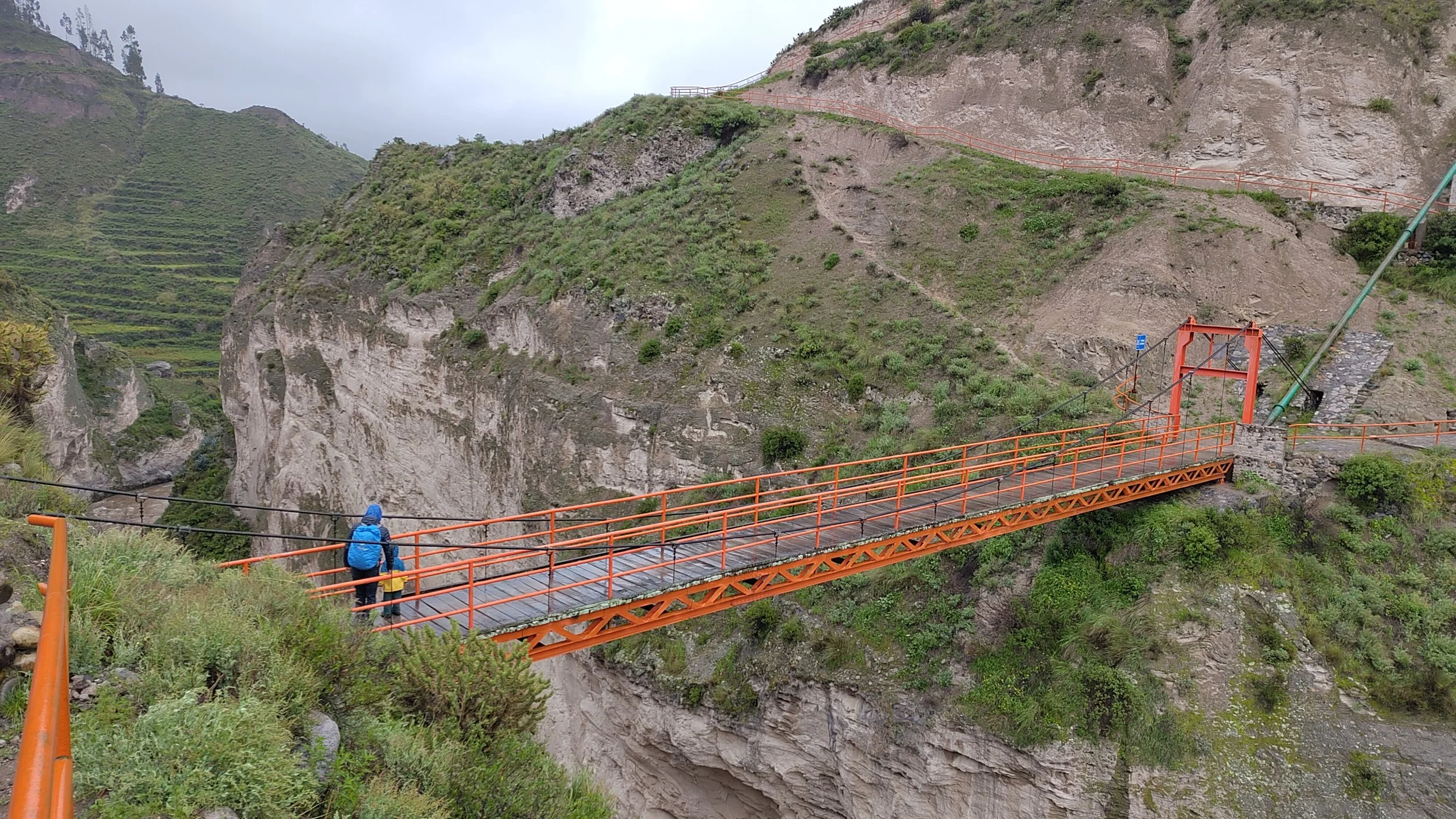 eine Hängebrücke in einer bergigen Landschaft mit grünen Hügeln und Felsen, eine Person mit blauen Jacke und Rucksack läuft auf der Brücke